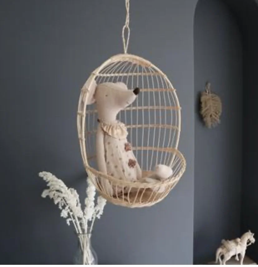 A teddy bear sitting inside a hanging wicker egg-shaped chair in a room with dark blue walls. There is a white vase with dried flowers on a table nearby and a small horse figurine on the floor.