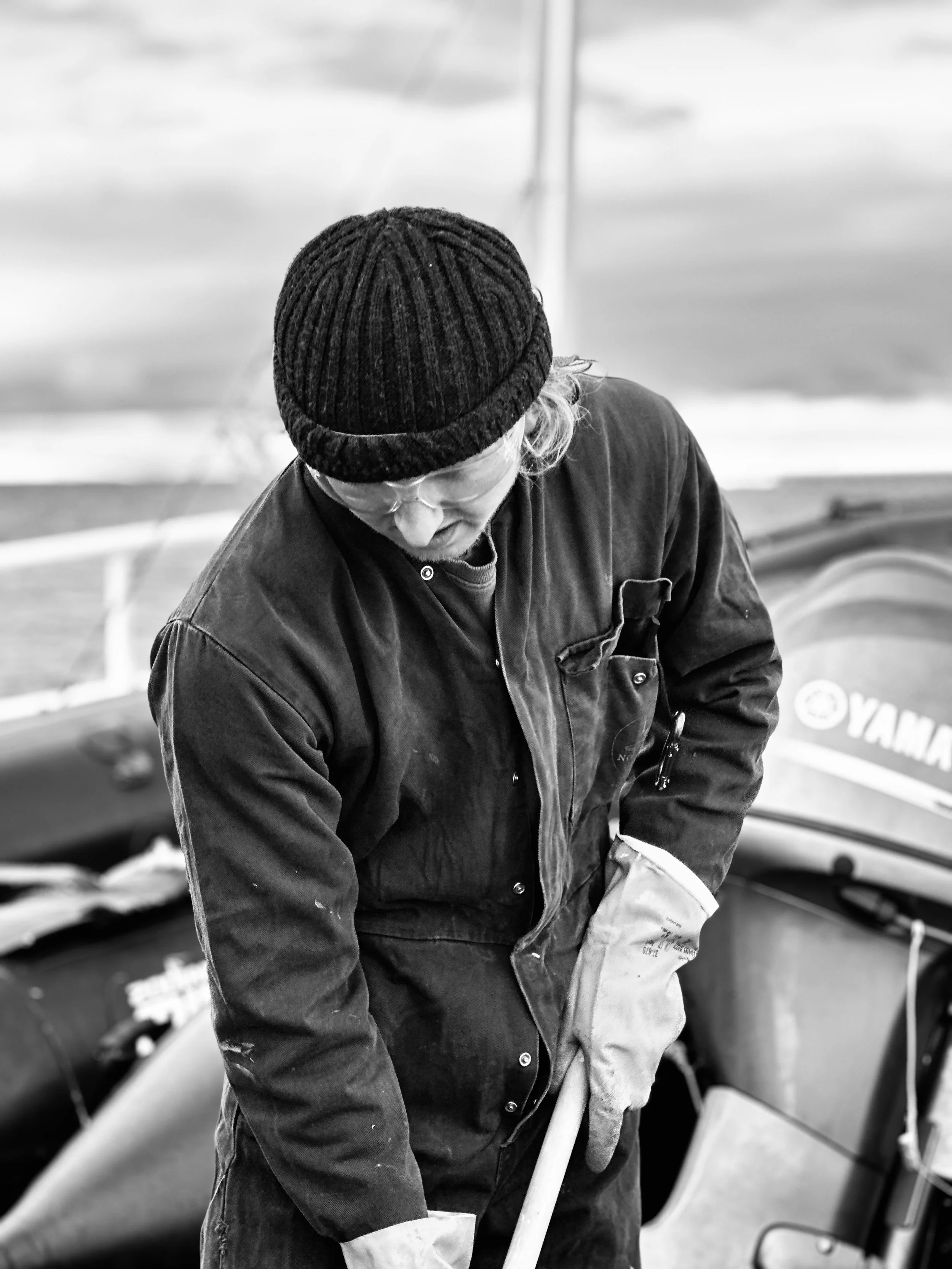 A man wearing glasses, a knit cap, and a dark jacket looking down while working on a boat, with a Yamaha outboard motor visible in the background.