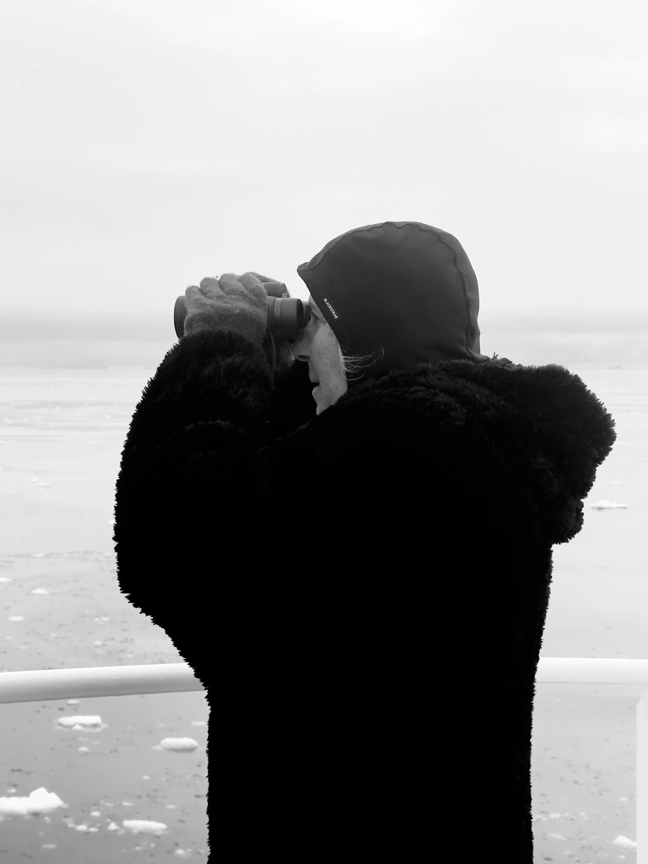 Person in a black furry coat and hood looking through binoculars at icy landscape