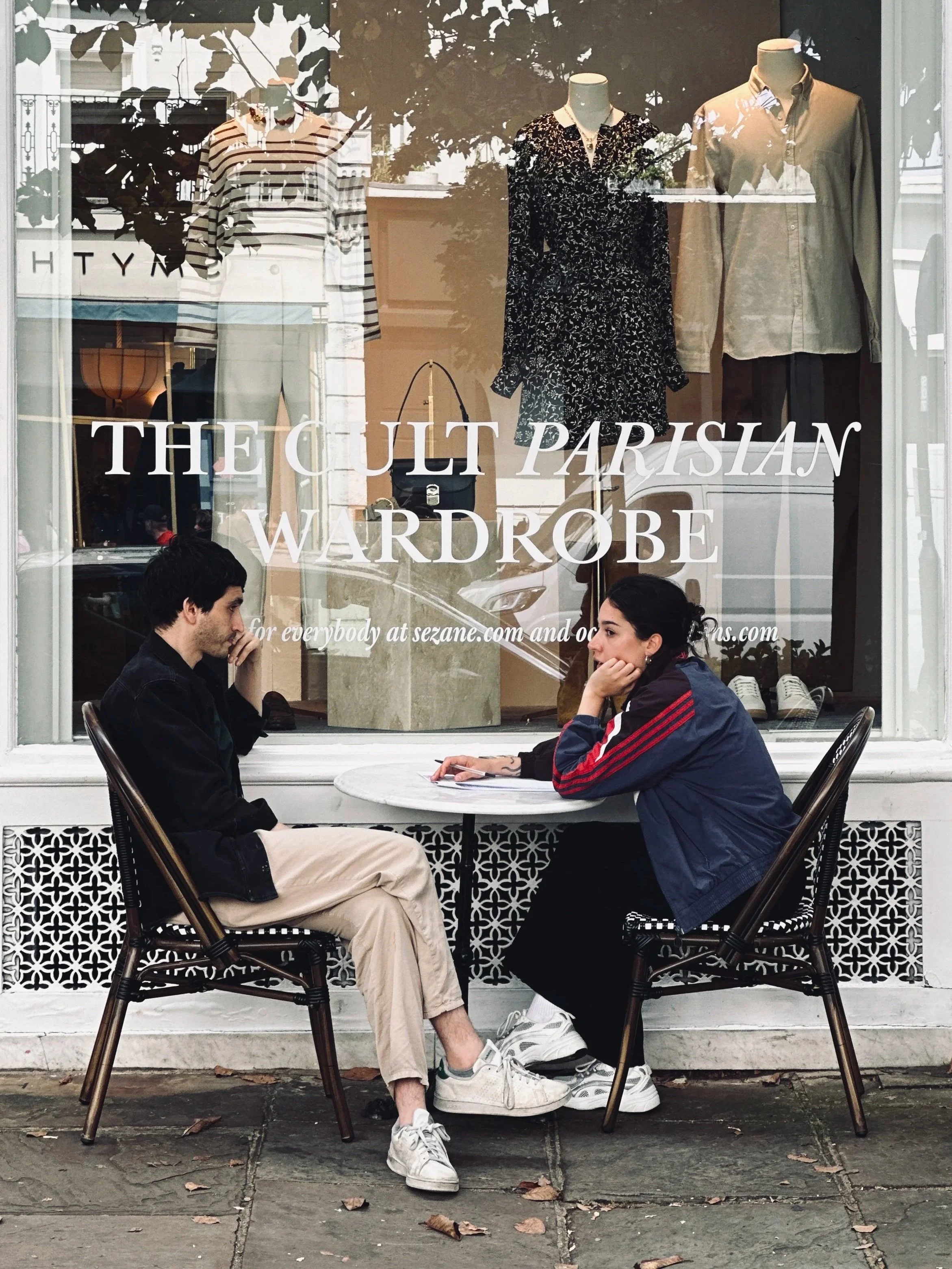 Two people at a bistro table outside the window of The Cult Parisian Wardrobe, a French fashion boutique