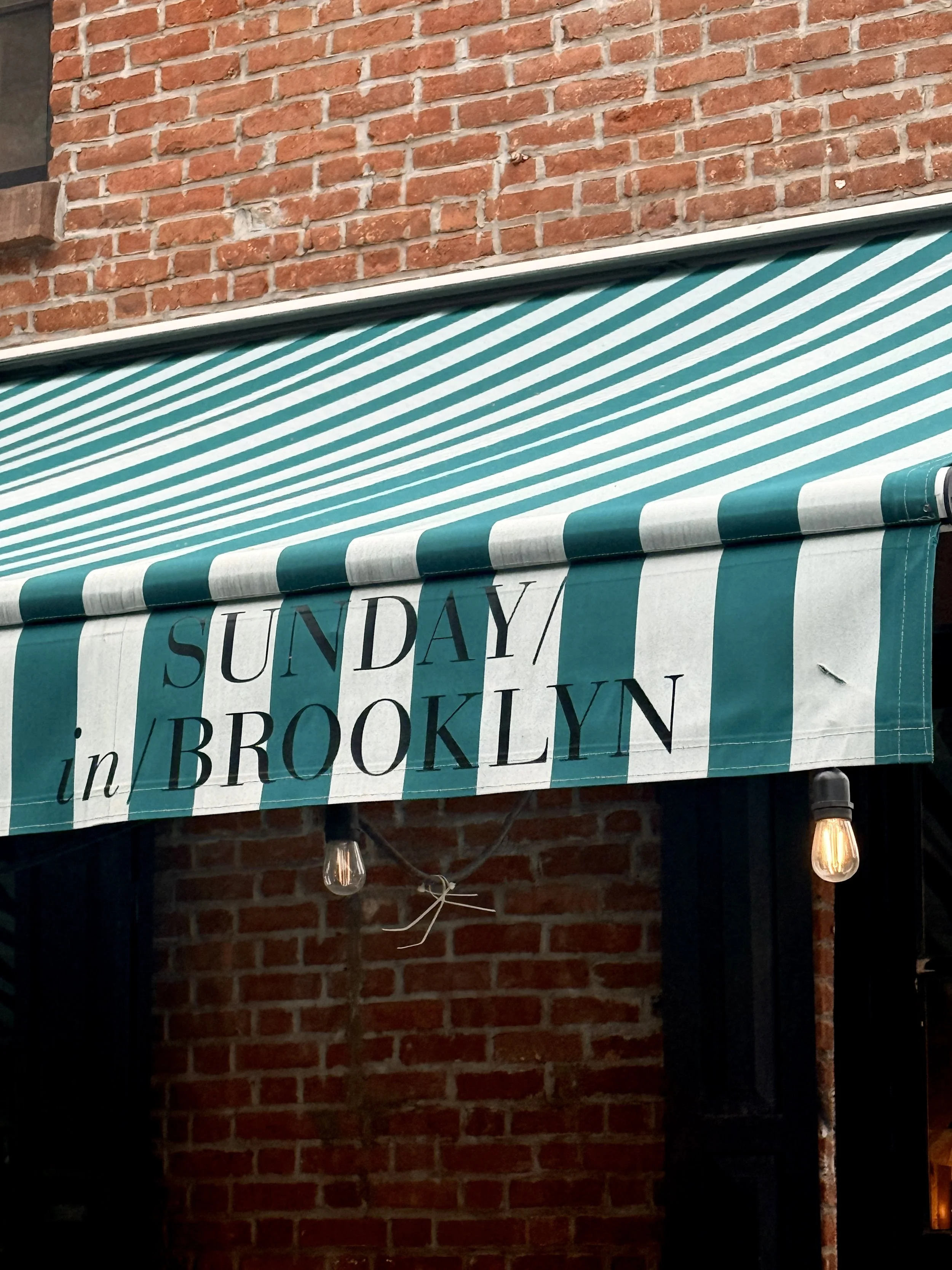Green-and-white striped awning with Sunday in Brooklyn lettering on a brick façade, Brooklyn