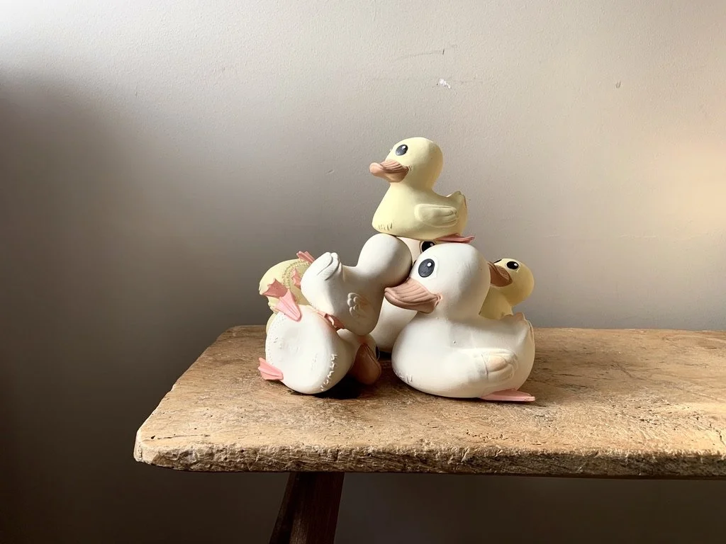 Five rubber duck toys stacked and placed on a wooden surface against a plain wall.