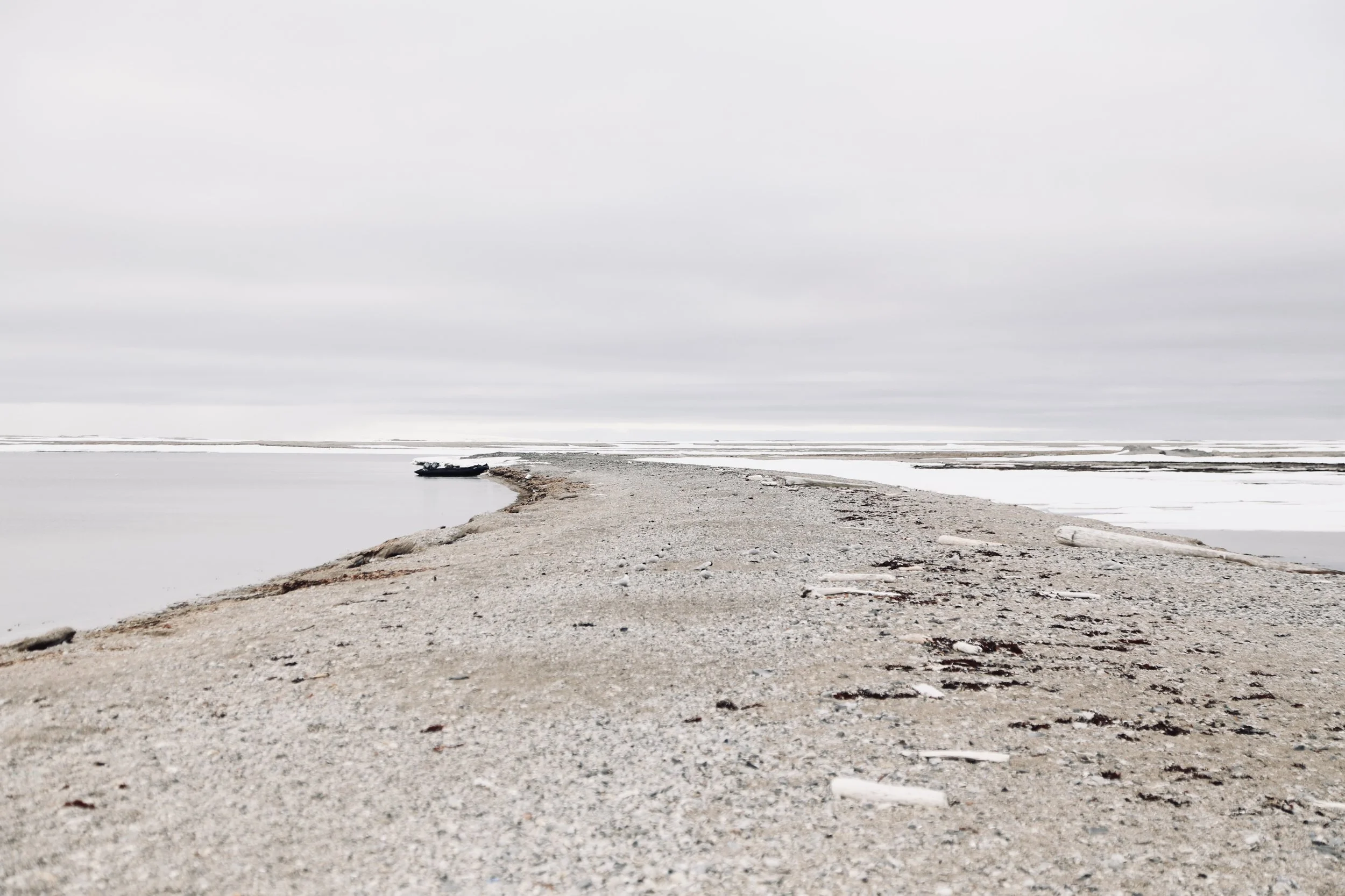 A narrow sand and dirt pathway extending into a body of water, with an overcast sky and few scattered pieces of driftwood on the shoreline.