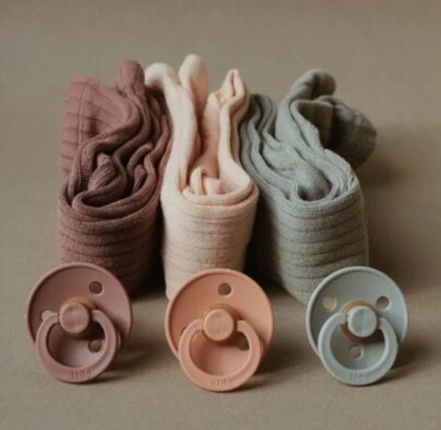 Three rolled-up baby socks in pink, cream, and gray, each with a matching pacifier in front.