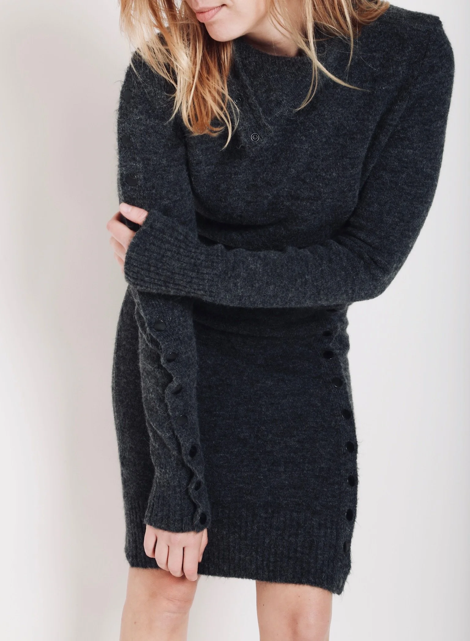 A woman with blonde hair wearing a dark gray, long-sleeved knit dress with button details on the sleeves and side, standing against a plain background.