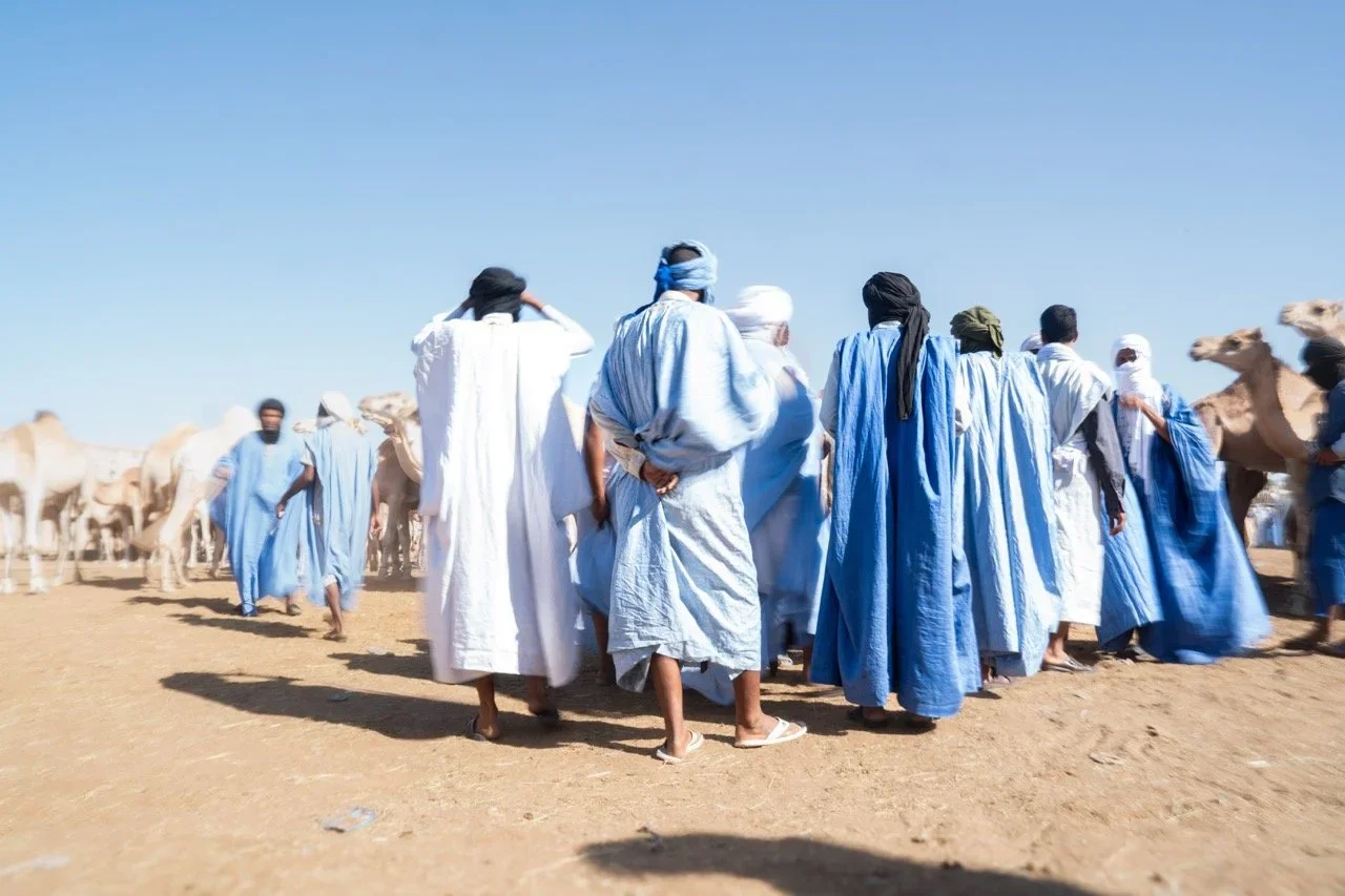 A group of people dressed in traditional white and blue robes walking across a sandy desert landscape, with camels on the sides under a clear blue sky.
