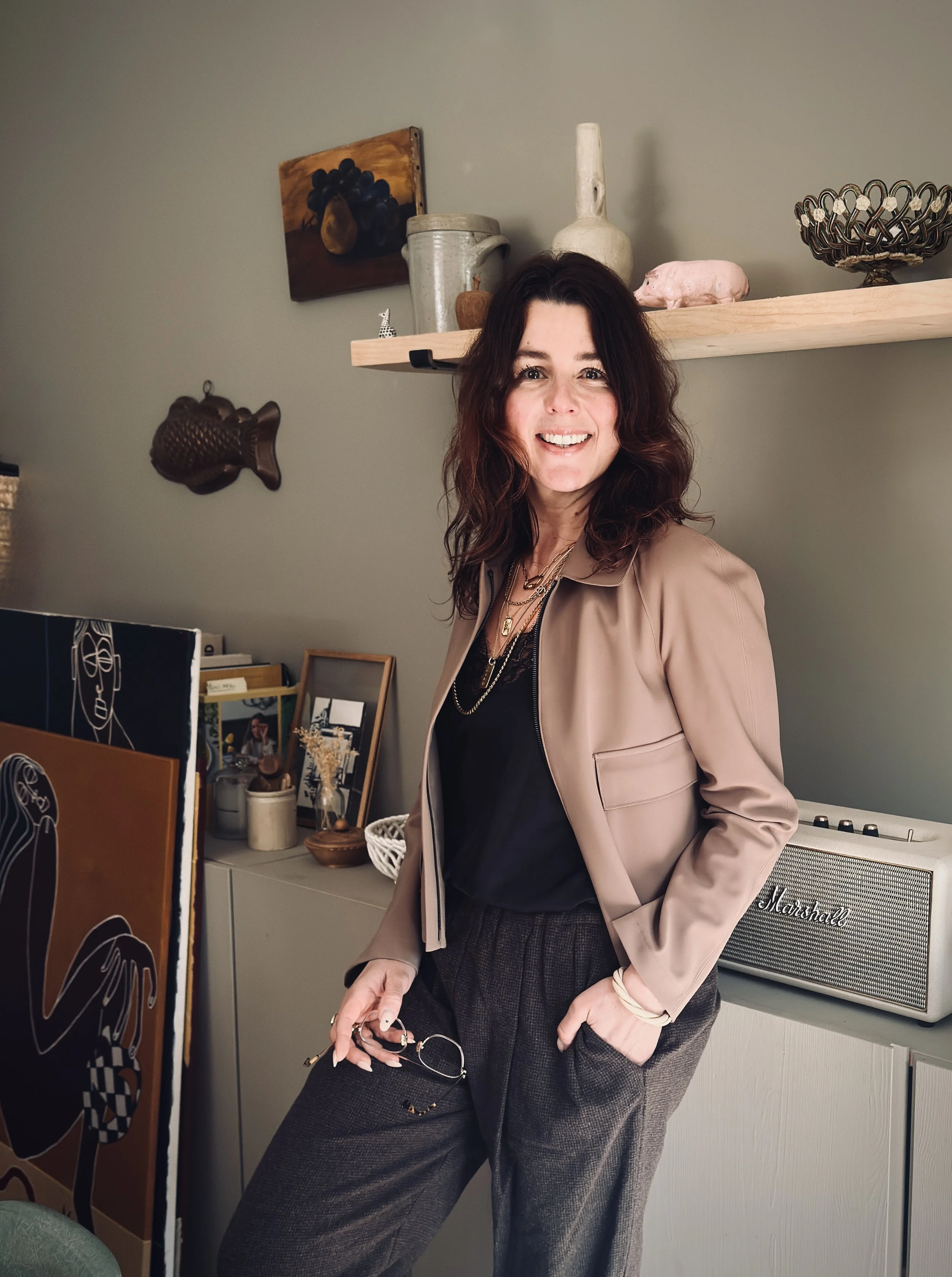 A woman with shoulder-length wavy dark hair smiling, wearing a beige blazer over a black top, standing in a room with decorative items and art on the wall.