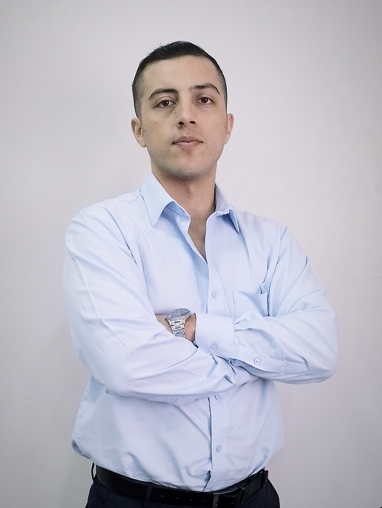 Hombre joven con camisa azul claro, de cabello corto, cruzado de brazos, con reloj en la muñeca izquierda, frente a fondo de pared blanca.