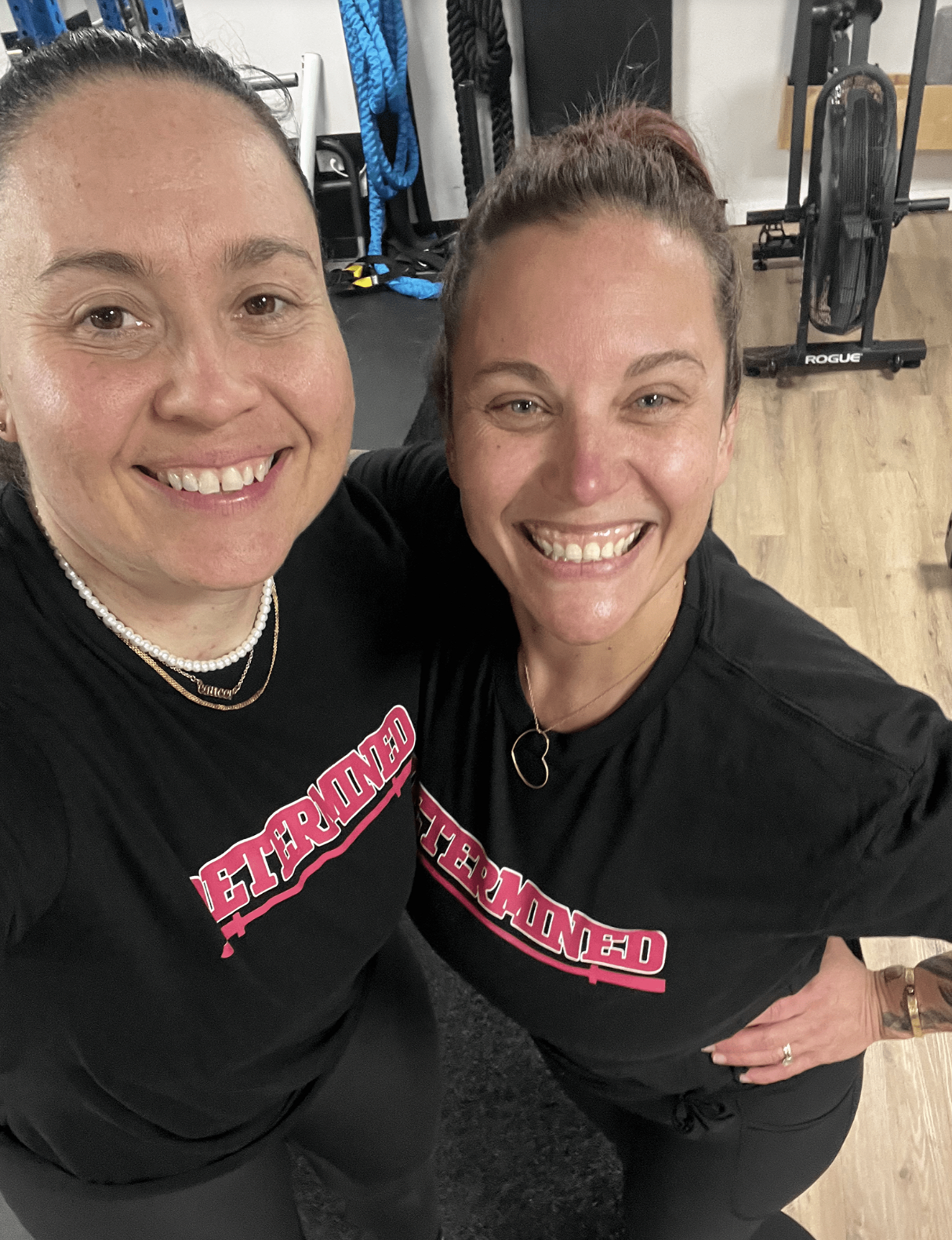 Two women smiling in a gym, wearing matching black shirts with pink and white text, surrounded by workout equipment.