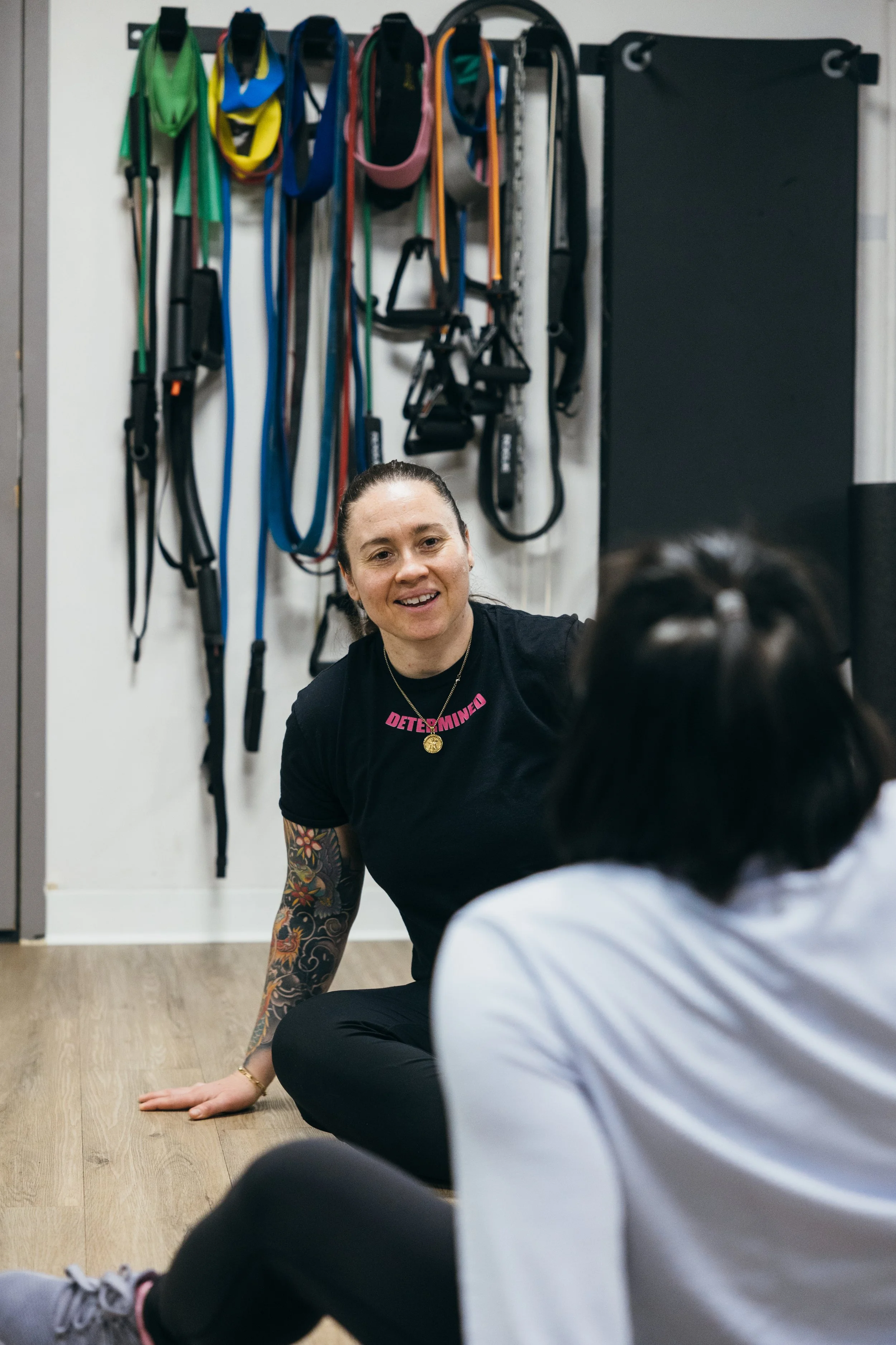 A woman with tattoos on her arm is sitting on the floor in a gym, engaging with a person in front of her. Behind her, there are workout resistance bands and straps hanging on a wall, and a black padded wall to the right.