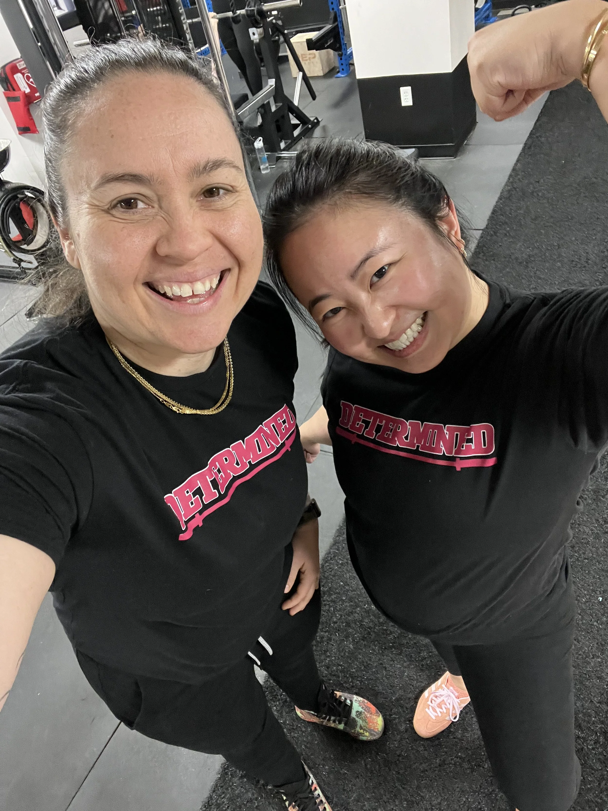 Two women in workout shirts with 'DETERMINED' written on them taking a selfie at the gym. One woman has dark hair tied back, and the other has brown hair, both showing big smiles.