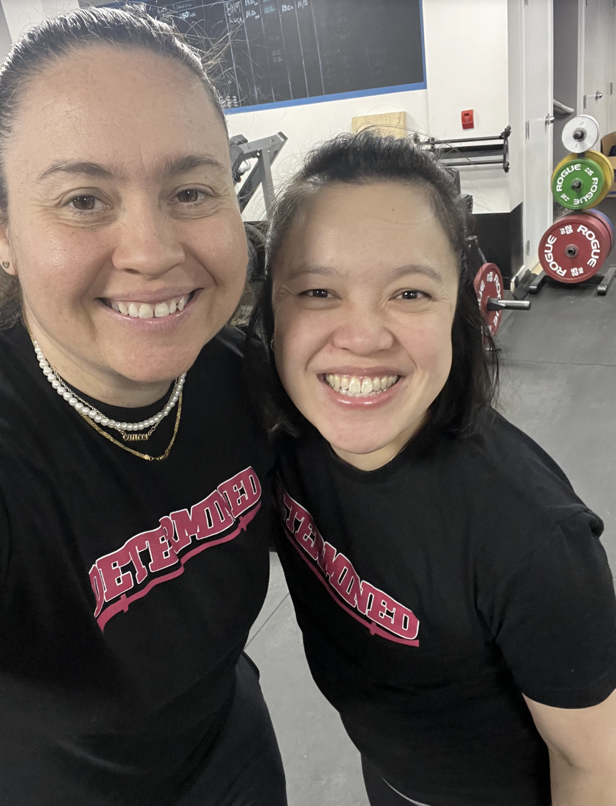 Two women smiling and taking a selfie in a gym with weightlifting equipment in the background. They are wearing black T-shirts with red and white text.