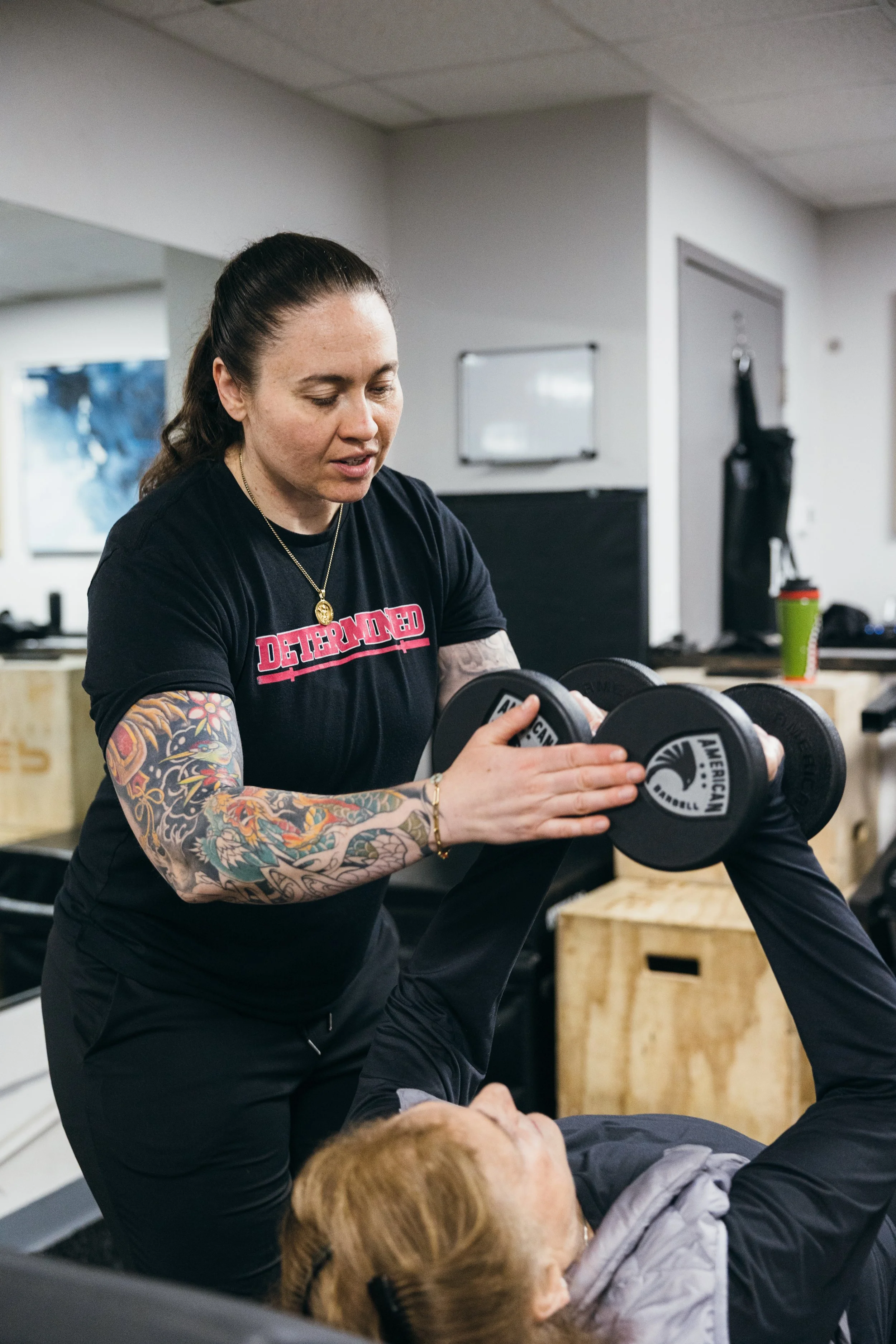 A woman with dark hair tied back, tattoos on her arms, and wearing a black shirt, is spotting a person during a workout, in a gym setting. The woman has a gold necklace and is holding adjustable dumbbells.