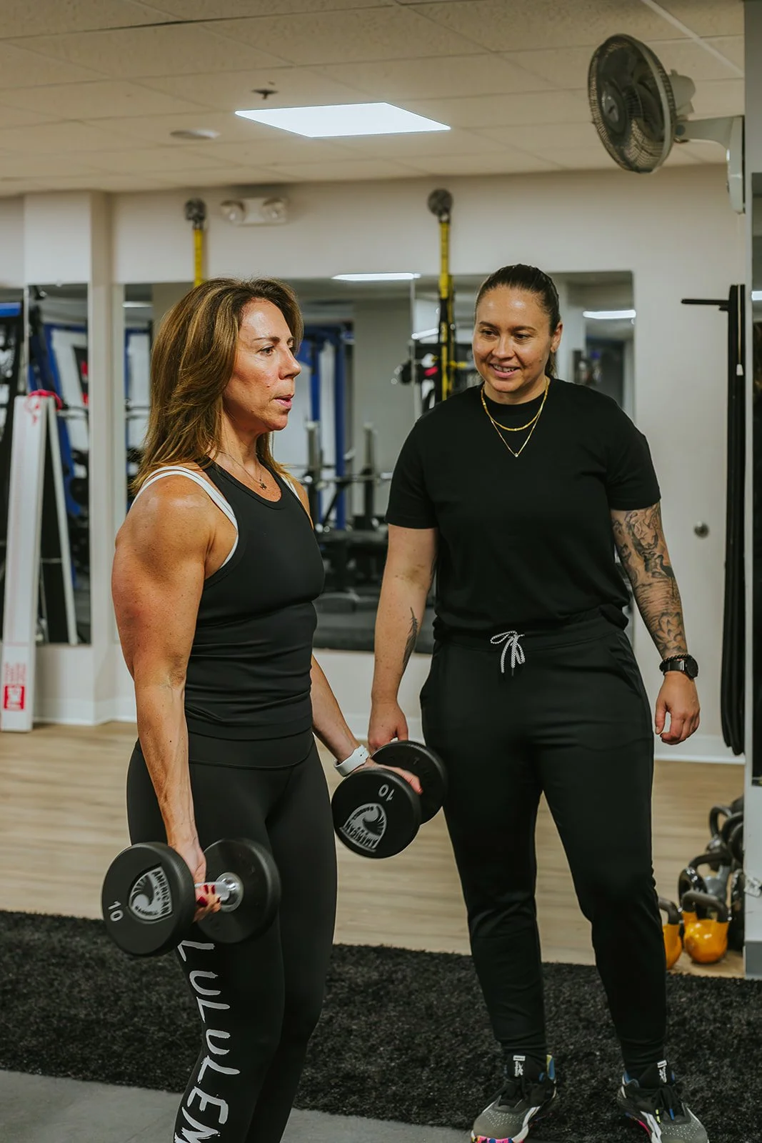 Two women in a gym, one lifts dumbbells while the other supervises or instructs.