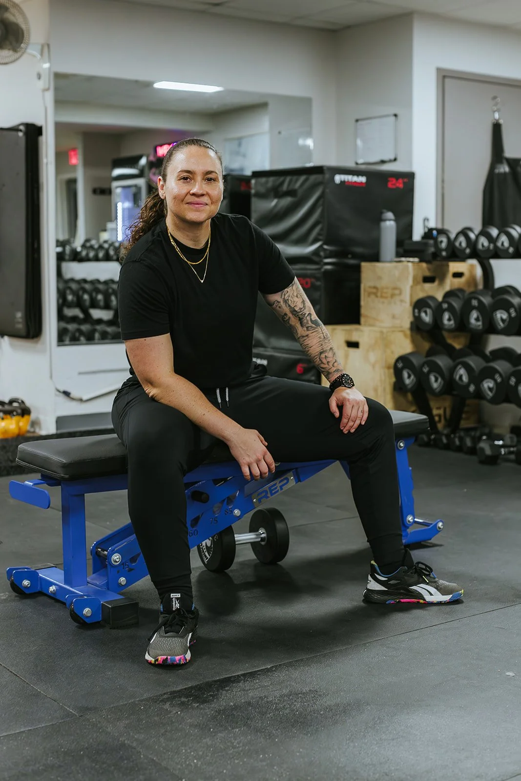 A woman sitting on a blue adjustable workout bench in a gym surrounded by dumbbells, kettlebells, and gym equipment.