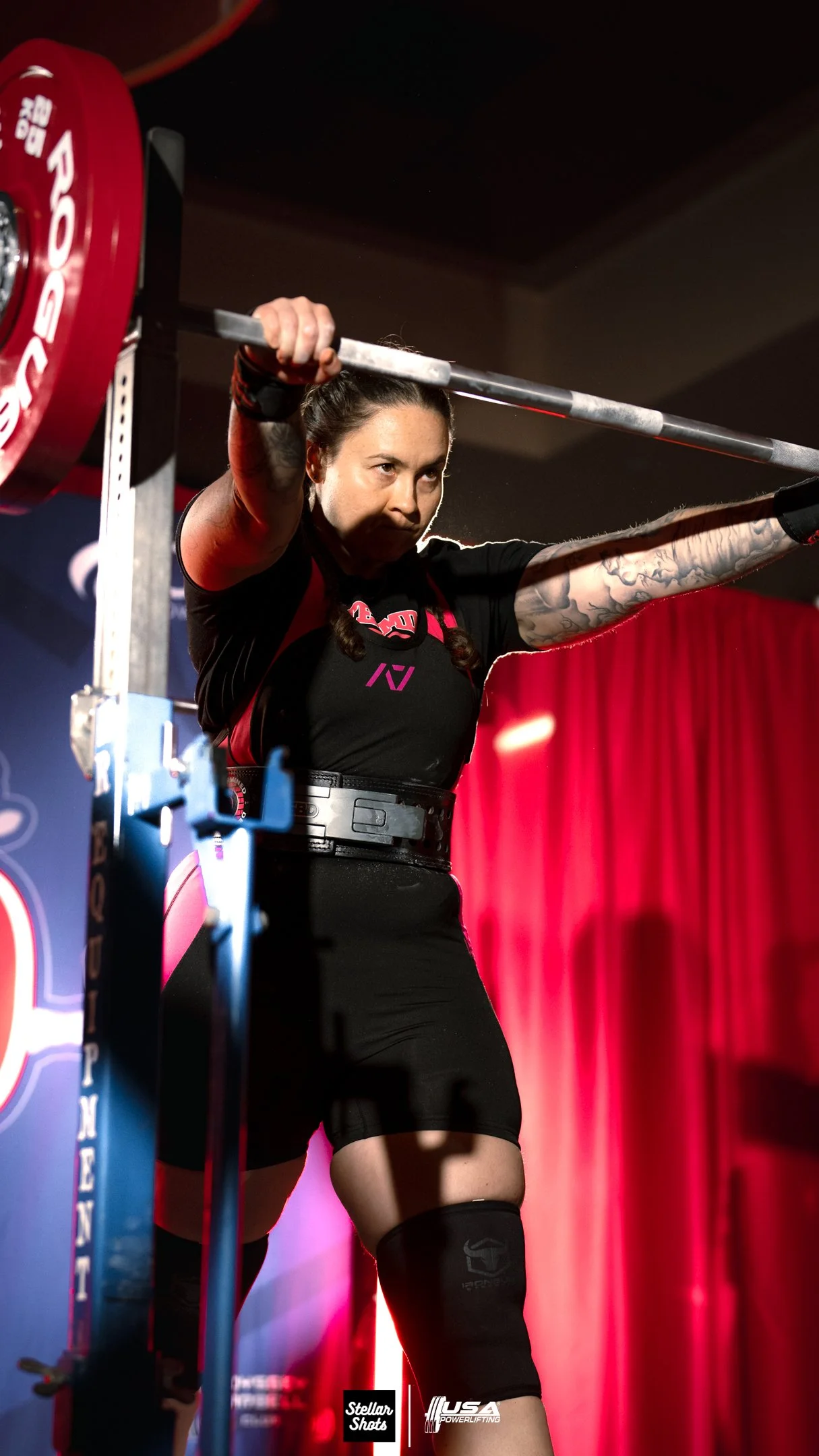 Female powerlifter preparing to lift a barbell, wearing a lifting belt, black shirt, and knee sleeves, in front of a red curtain at a competition.