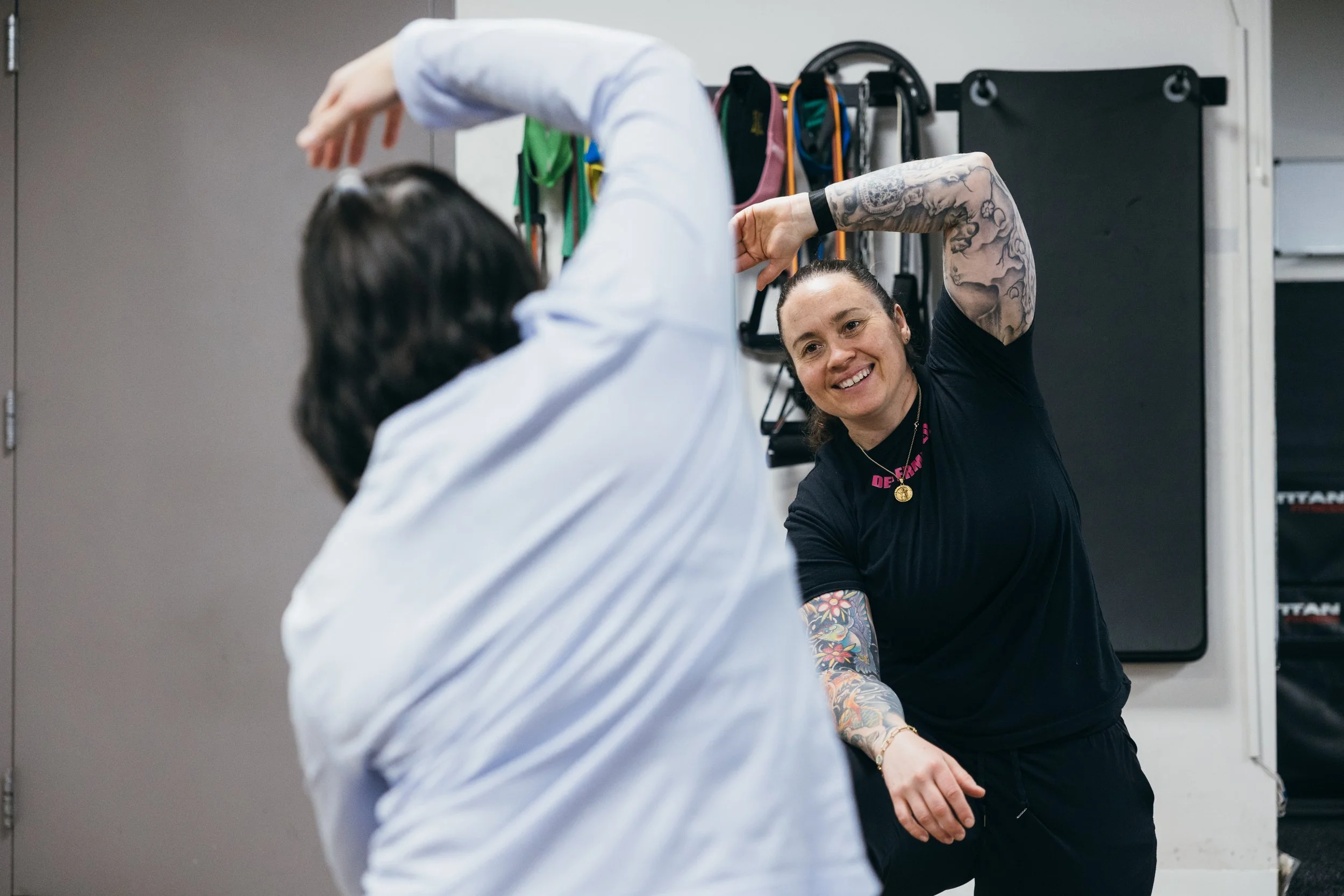 A woman with tattoos smiling and raising her arm during a workout or self-defense class with another person in a gym or studio.