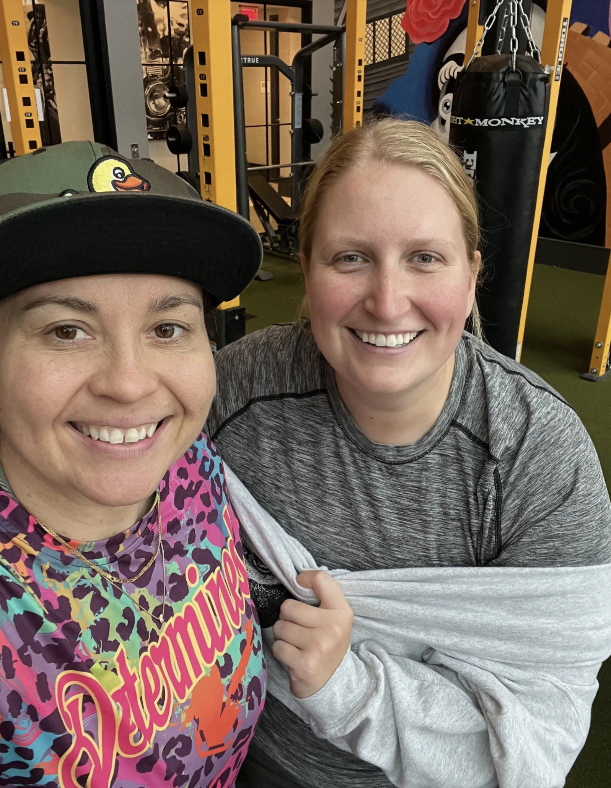 Two smiling women taking a selfie in a gym, surrounded by workout equipment, including a punching bag and weight racks.