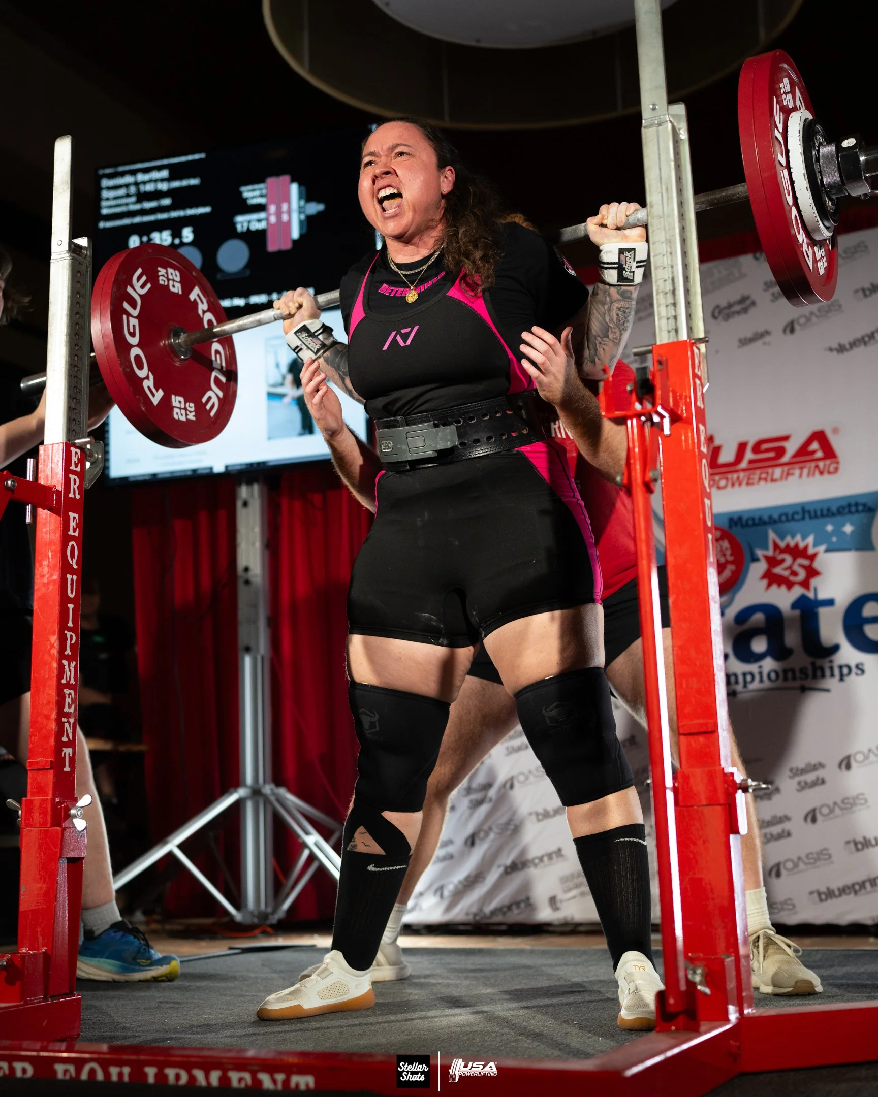 A woman performing a weightlifting squat with a barbell loaded with red weights at a powerlifting competition. She is wearing a black and pink sports outfit, knee sleeves, and white shoes, and appears to be exerting effort.