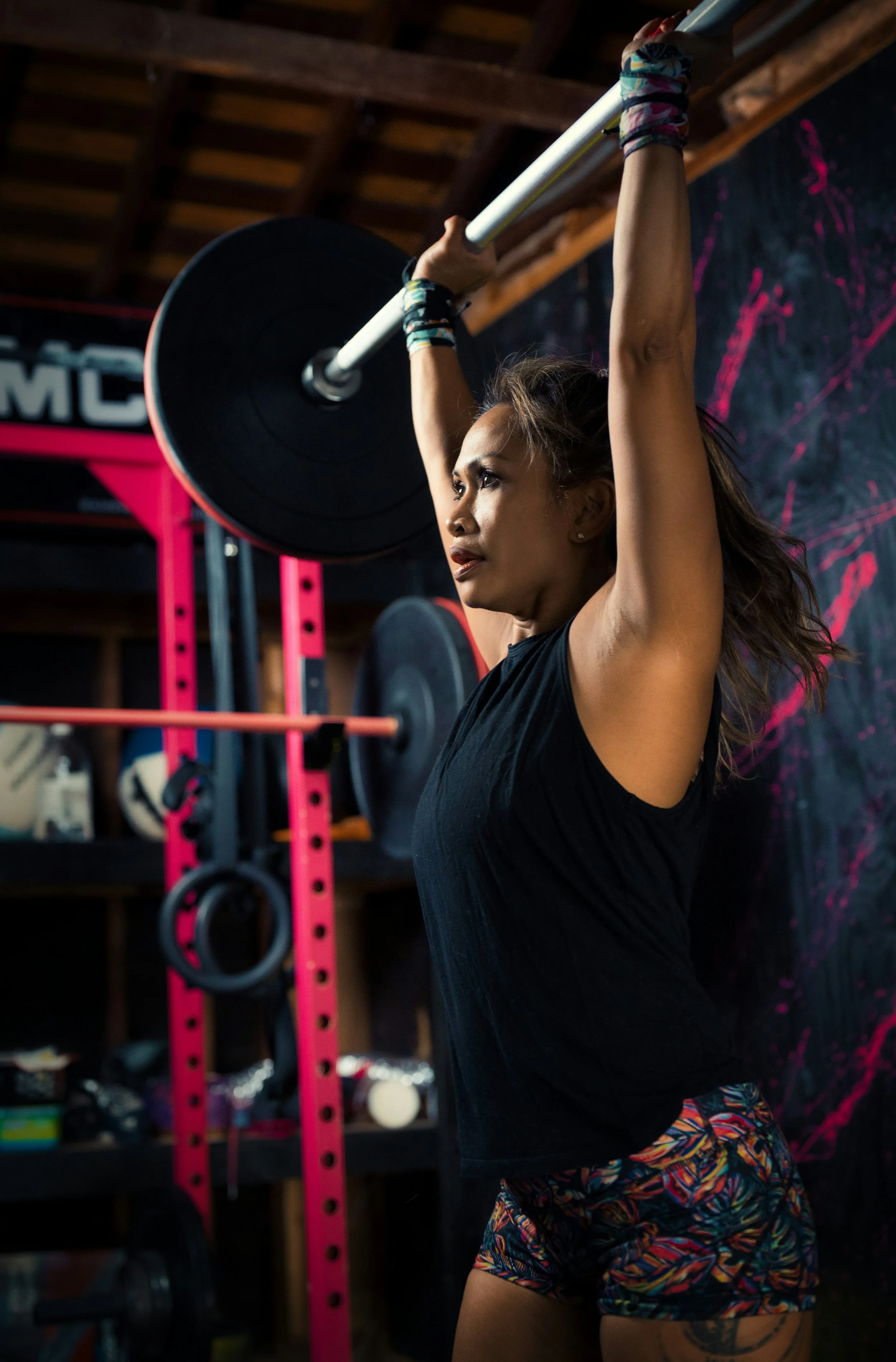 A woman is lifting a barbell in a gym. She is wearing a black workout top, colorful shorts, and has patterned wristbands. The gym background includes weight racks and a chalkboard with pink writing.