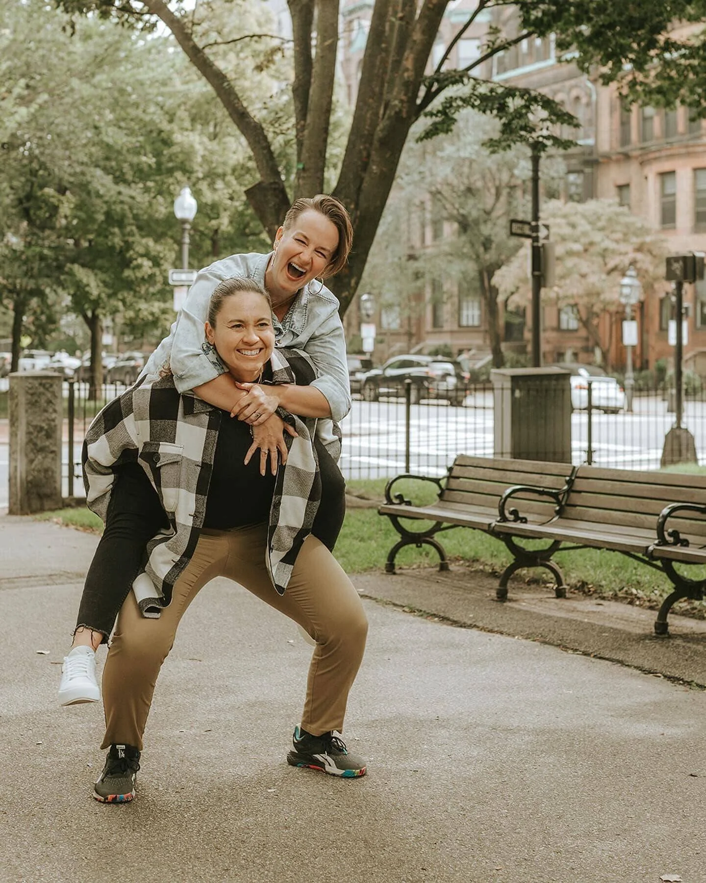 Two women smiling and having fun outdoors in a park, with one woman giving the other a piggyback ride, surrounded by trees and benches.