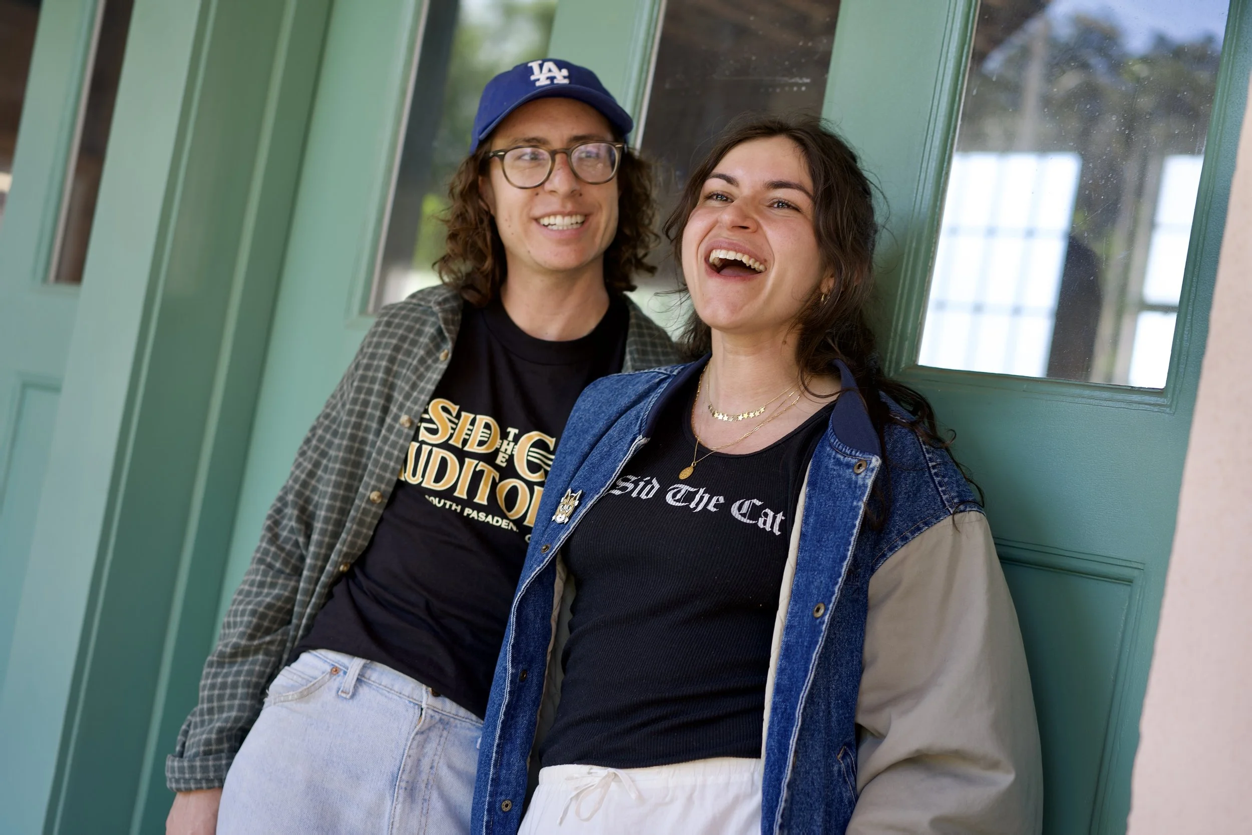 Two young women leaning against a green wall, smiling and laughing, dressed casually with one wearing a blue cap and glasses, the other with long dark hair, wearing denim jacket and black T-shirt.