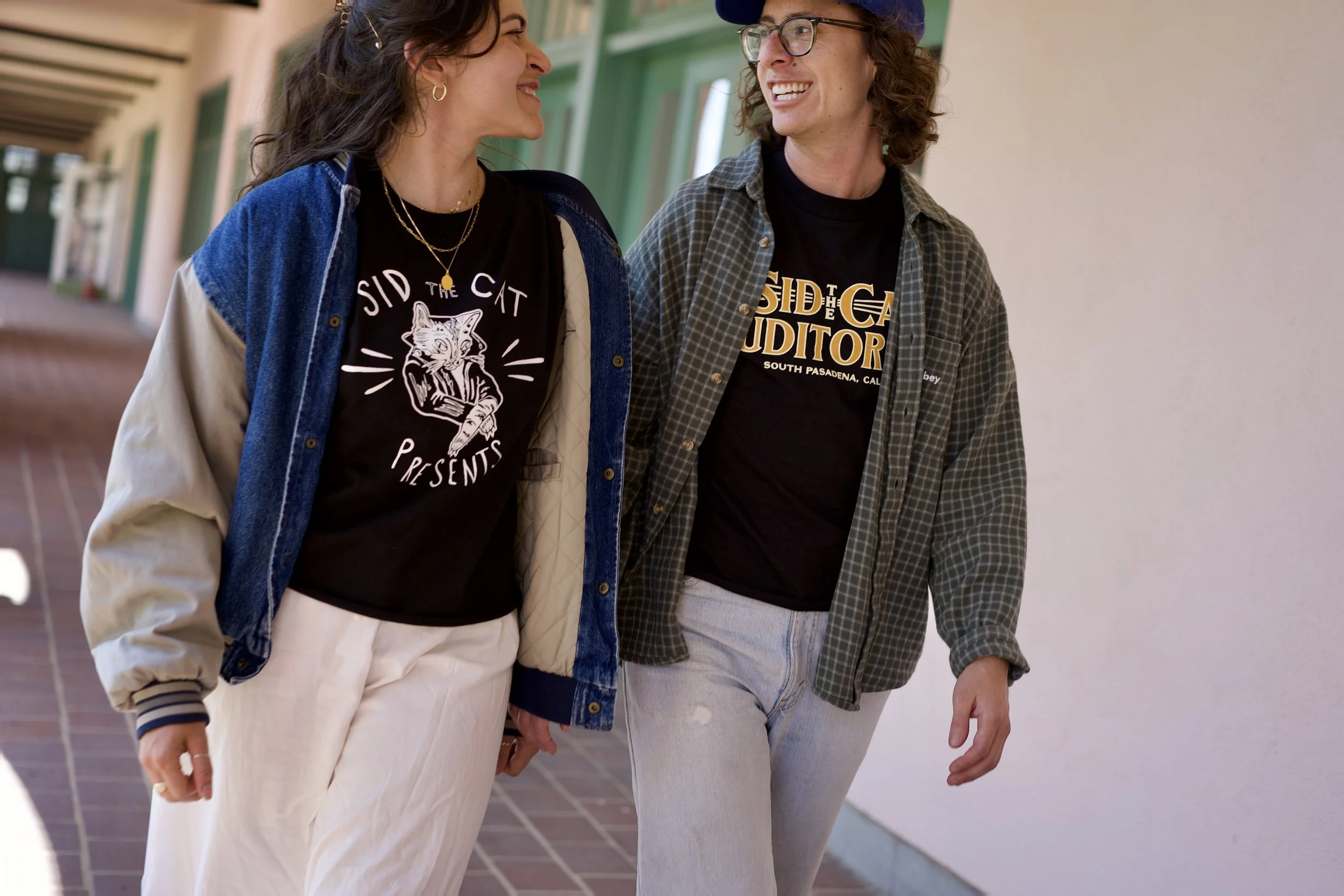 Two women walking and talking outdoors, smiling at each other. One woman has dark hair and is wearing a black T-shirt with a cat graphic, a blue and beige jacket, and cream pants. The other woman has curly hair, glasses, and is wearing a black T-shirt with yellow text, a plaid shirt, and light-colored pants.