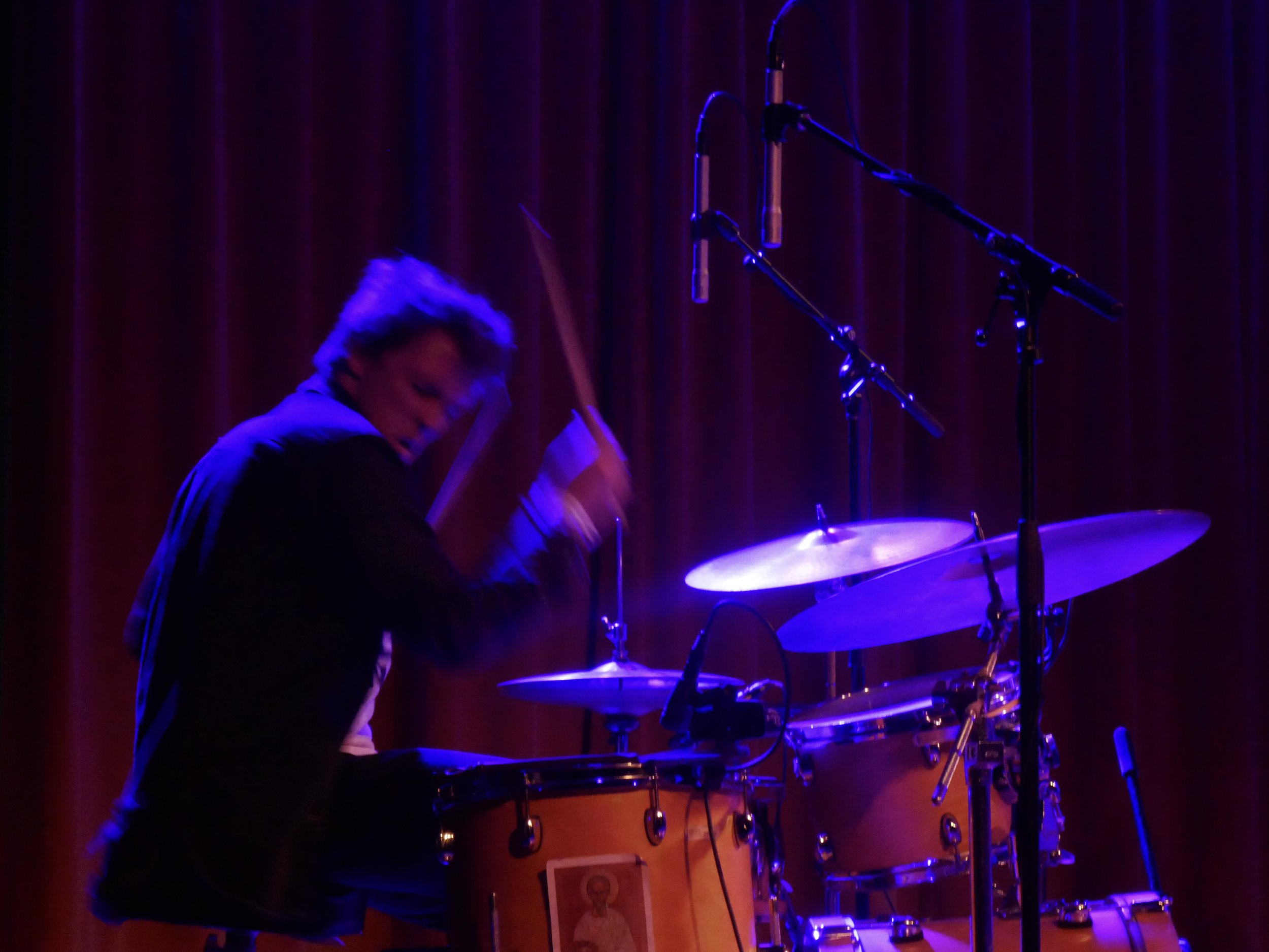 A person playing drums on stage with purple and blue lighting, standing behind a drum set with microphones overhead.