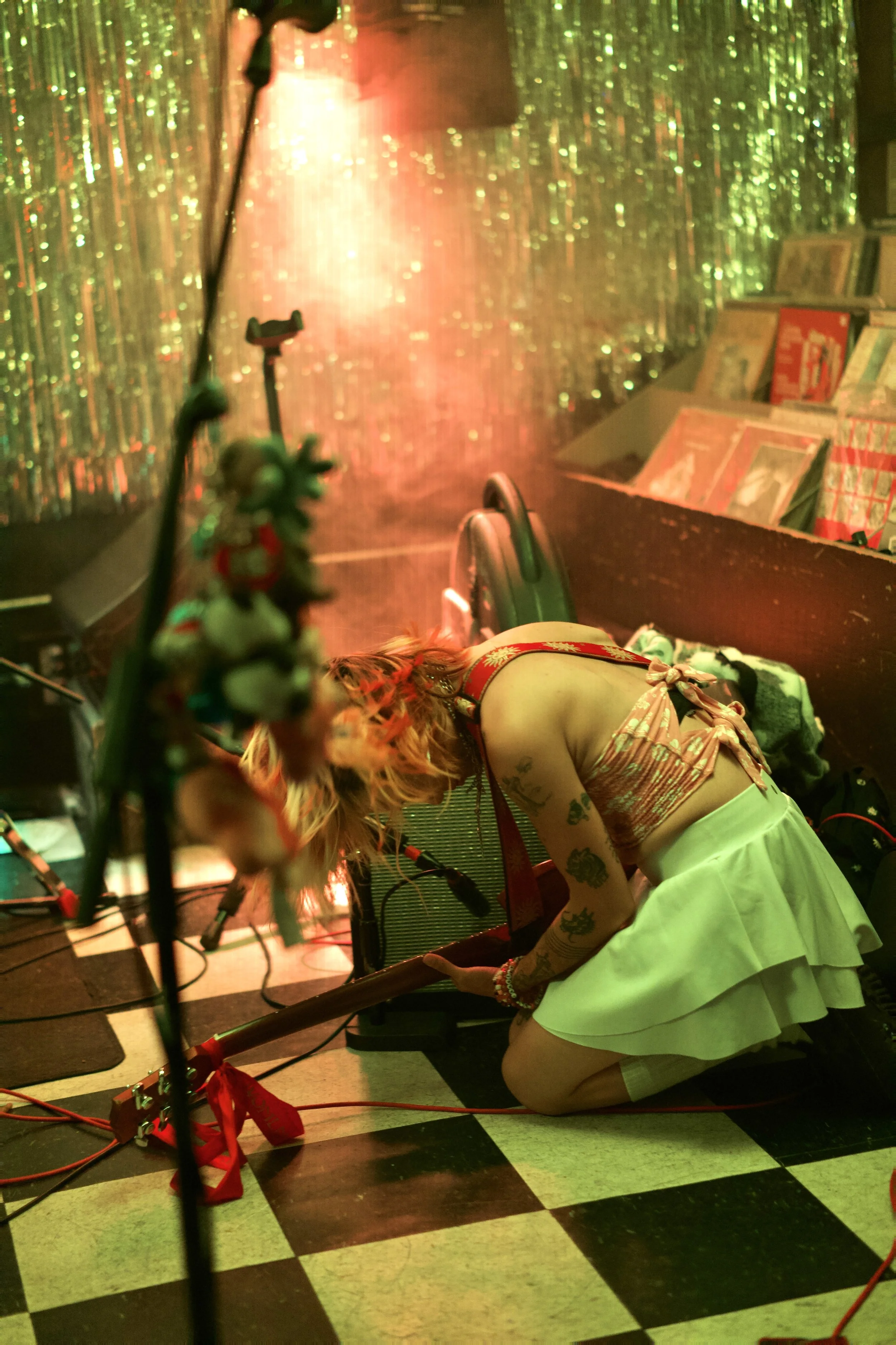 A woman with red hair, tattoos, and light skin, dressed in a vintage-style outfit with a pink floral top and white skirt, is kneeling on a checkered floor in a dimly lit room with a glittery backdrop. She is focused on a guitar amplifier, possibly adjusting it, and is surrounded by music equipment. The scene is decorated with books on a shelf and a blurred figure in the foreground.