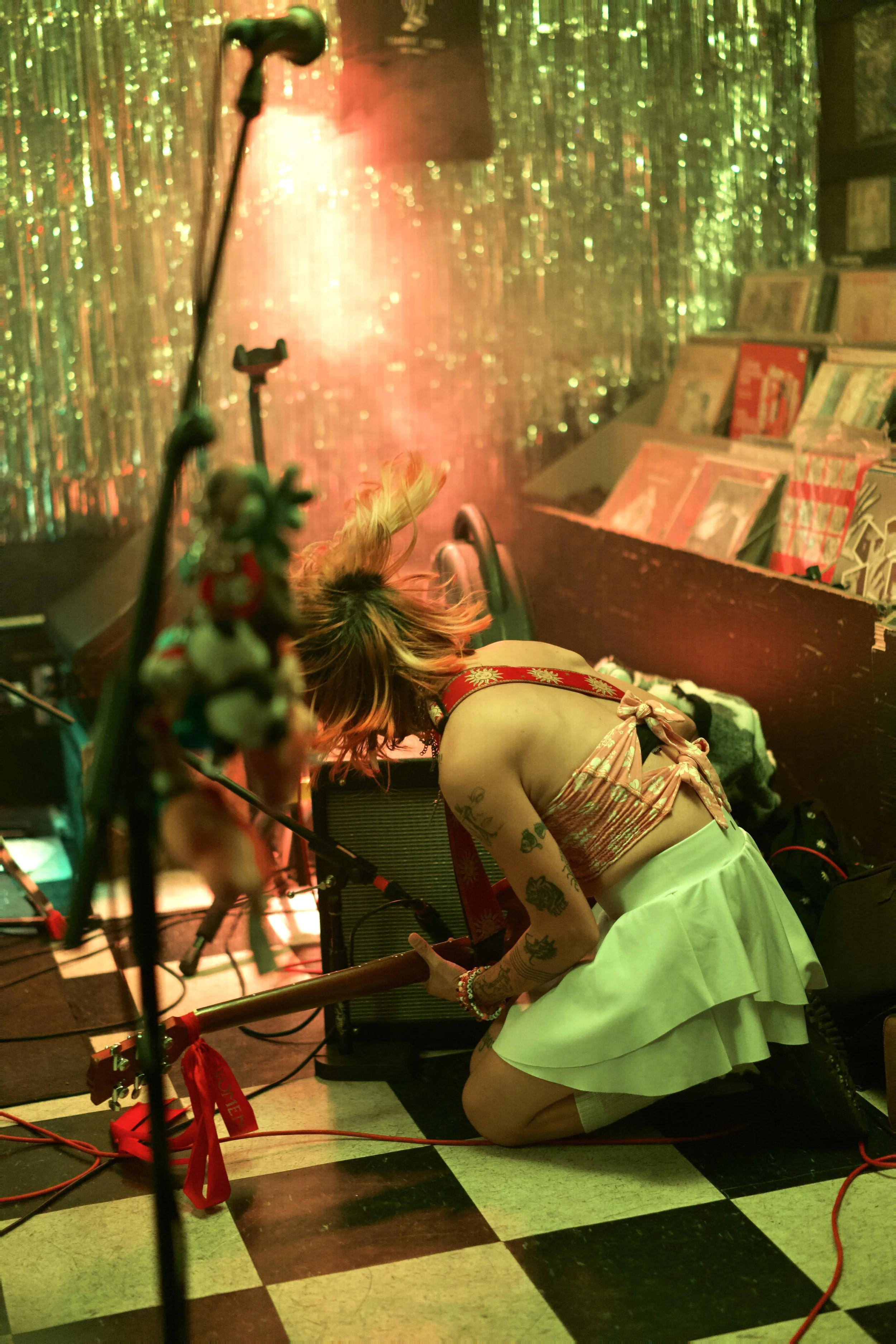 A woman with tattoos, wearing a pink top and a white skirt, kneeling on a black and white checkered floor. She is surrounded by electronic equipment and musical instruments, in front of a shimmering, colorful background.