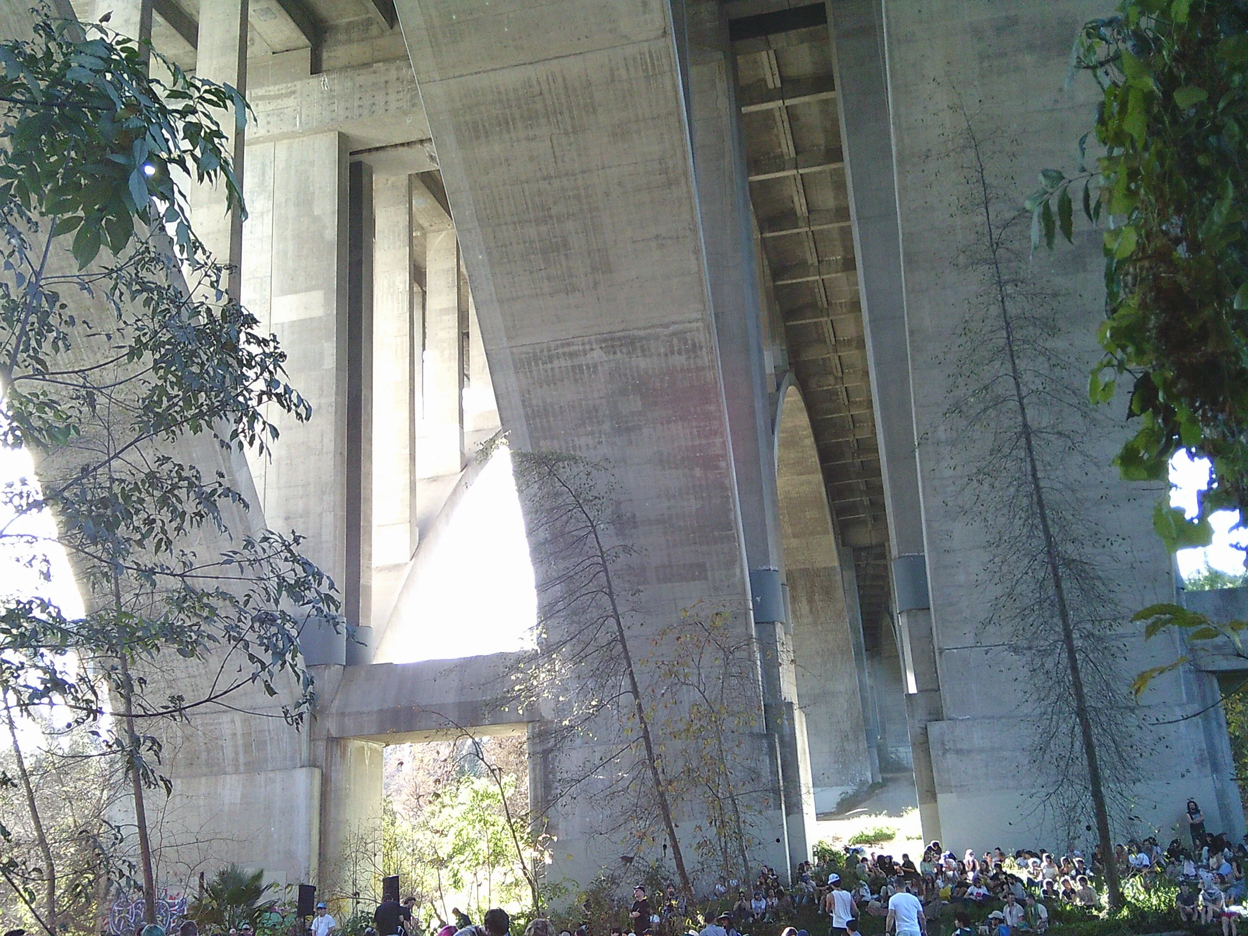 A large concrete building with multiple levels and open areas, surrounded by trees and plants. A crowd of people is gathered at the bottom right corner.