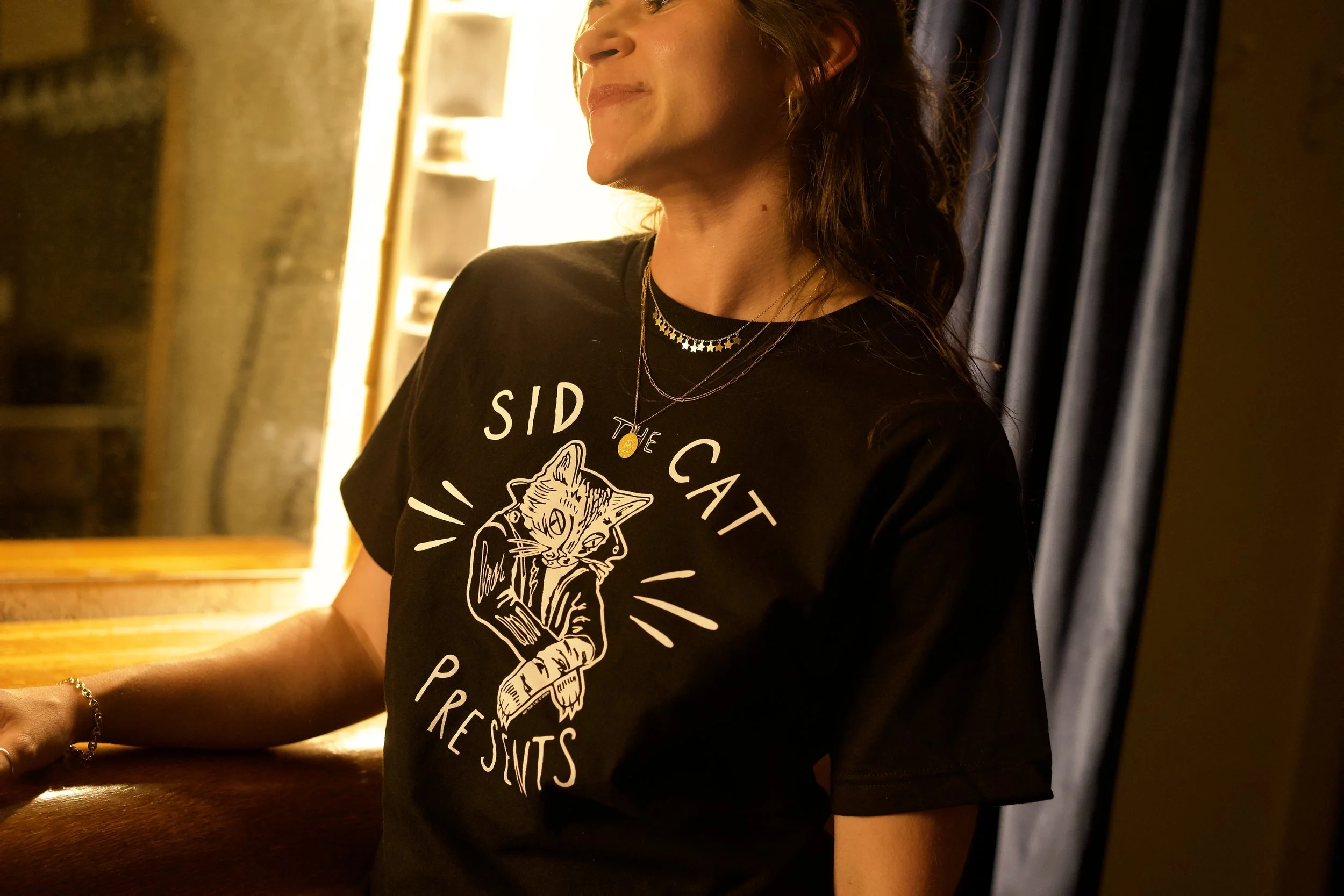 A woman wearing a black t-shirt with a graphic of a cat and the words 'Side Cat Presents' standing in a warmly lit room with dark curtains and a wooden surface.