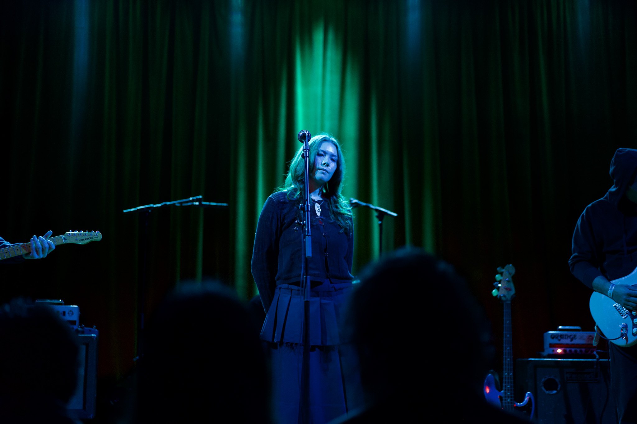 A woman performing on stage with closed eyes, surrounded by green stage lights, with musicians and musical equipment visible around her.