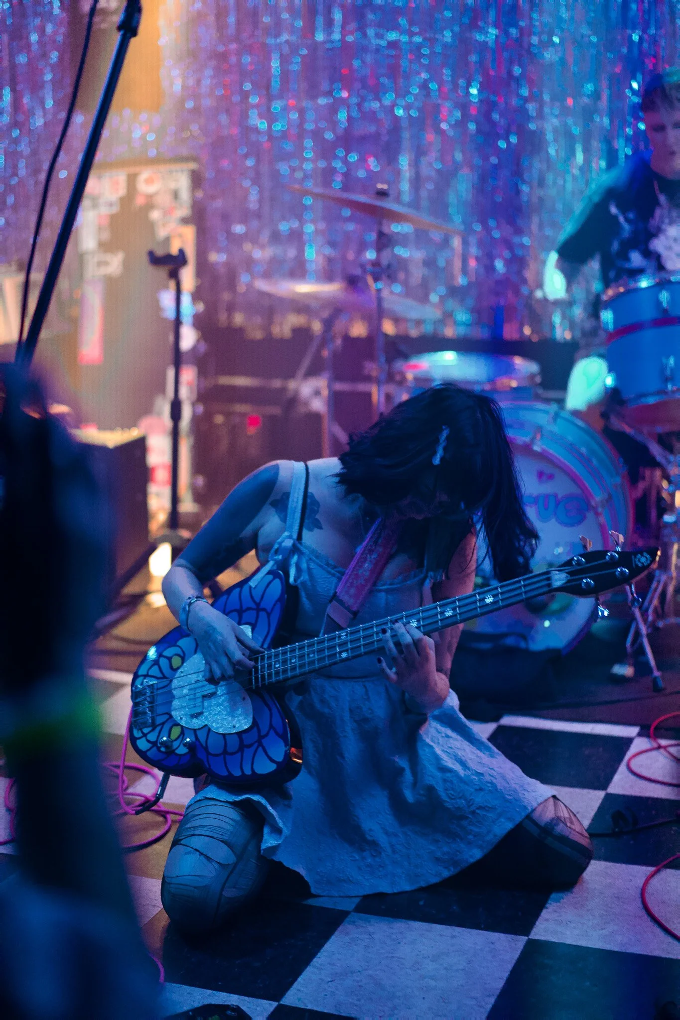 A woman kneeling on the floor playing an electric guitar with a large butterfly graphic on it, in a dimly lit stage with a blue and purple sparkle background and drums behind her.