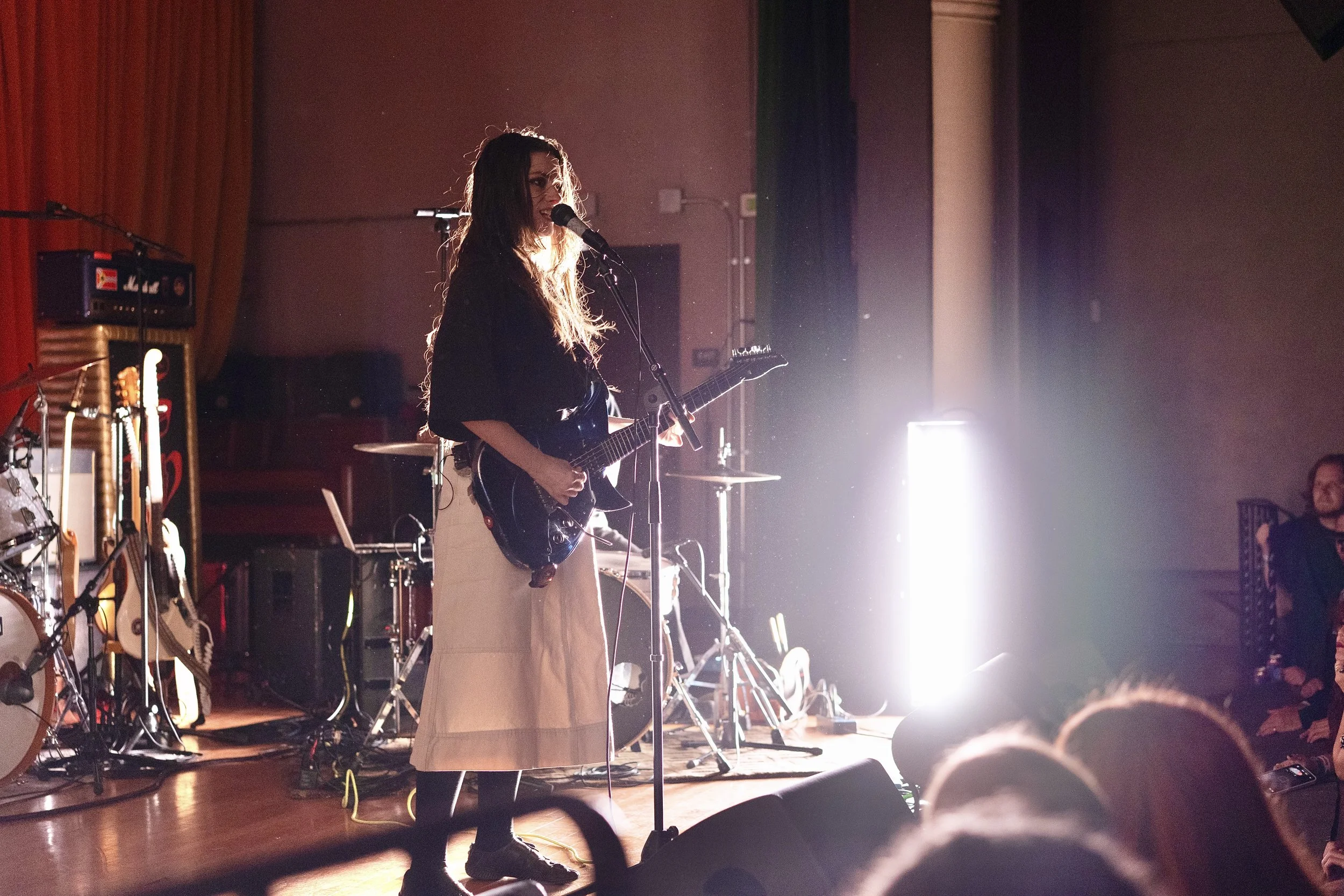 A woman with long hair playing an electric guitar and singing into a microphone on stage during a performance, with stage lights shining behind her.