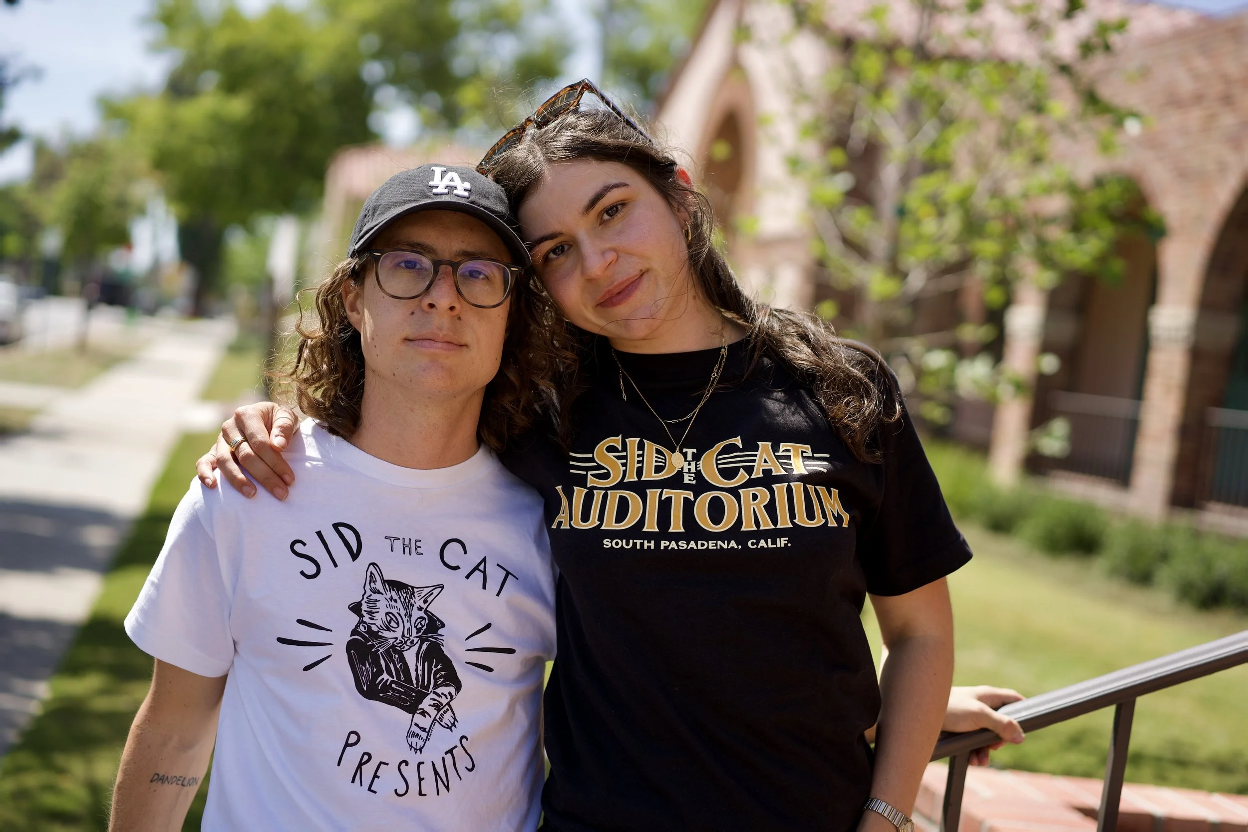 Two women stand close together outdoors, smiling at the camera. The woman on the left has glasses, a black LA Dodgers cap, and a white T-shirt with a cat illustration. The woman on the right has long, curly hair, sunglasses on her head, and a black T-shirt with yellow and white text. They are positioned on a sidewalk with green trees in the background, and a brick building is partially visible.