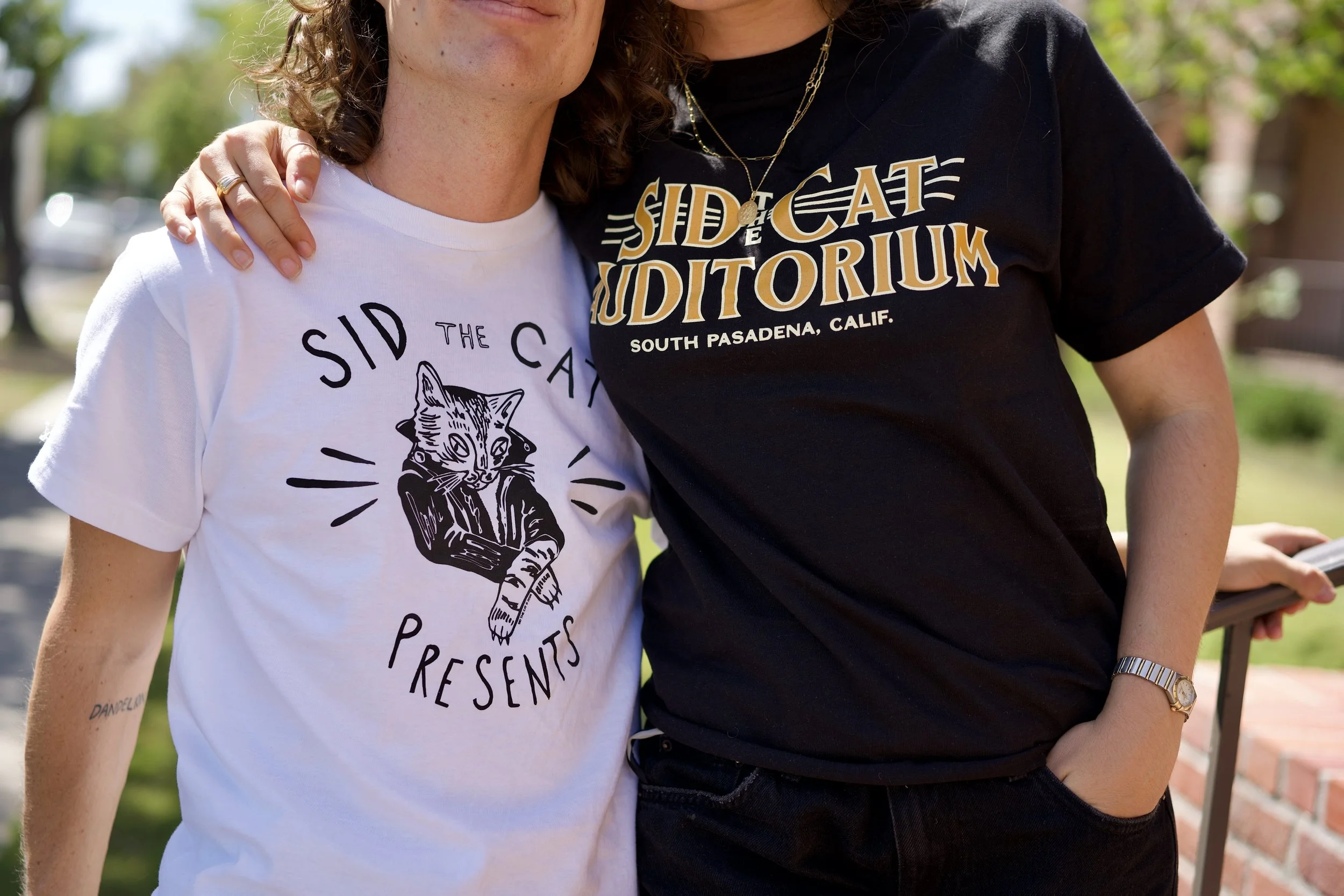 Close-up of two women standing outdoors, one wearing a white t-shirt with a cat graphic and the other wearing a black t-shirt with text, with green trees in the background.
