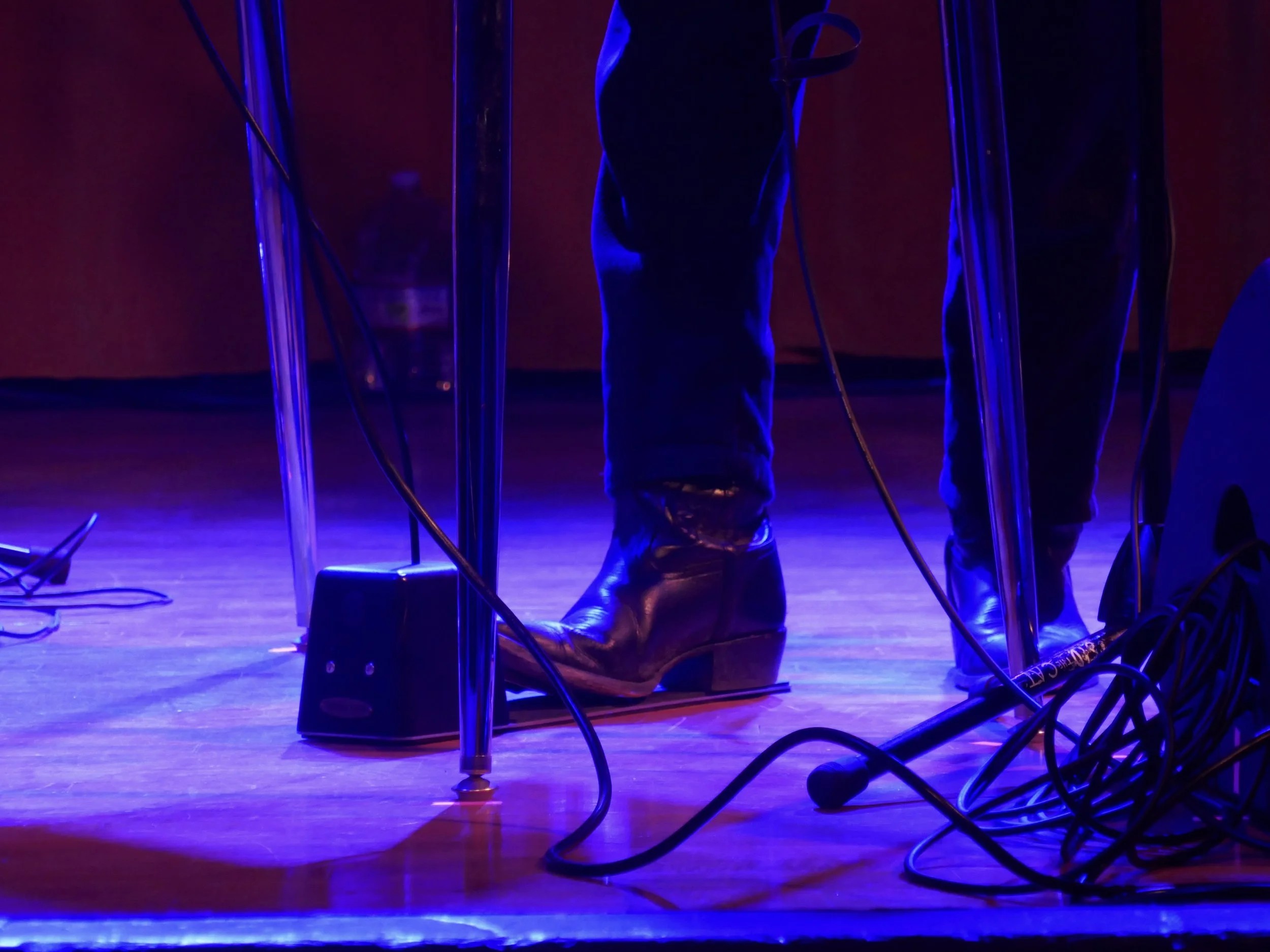 A person in jeans and a dark shirt is kneeling on a stage, surrounded by microphones, music stands, and cables, with purple and red stage lighting.