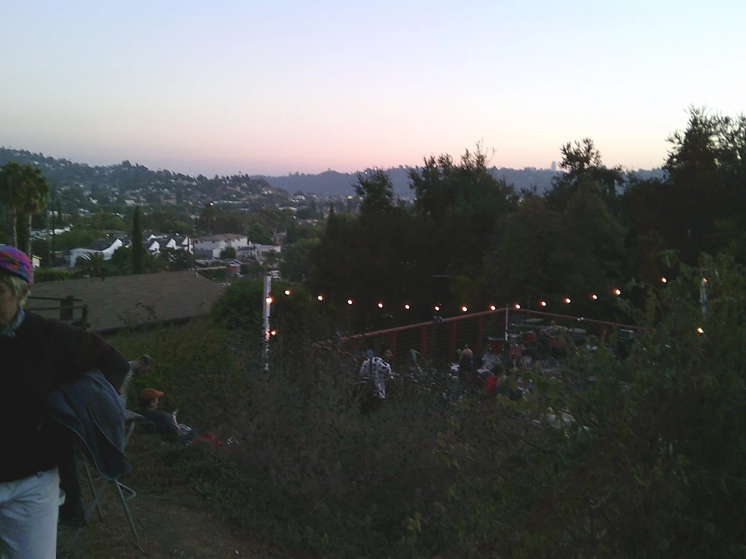 People gathered outdoors during sunset with trees and hillside in the background, some weathered string lights hanging, and a person partially visible on the left side wearing a dark jacket and light-colored pants.