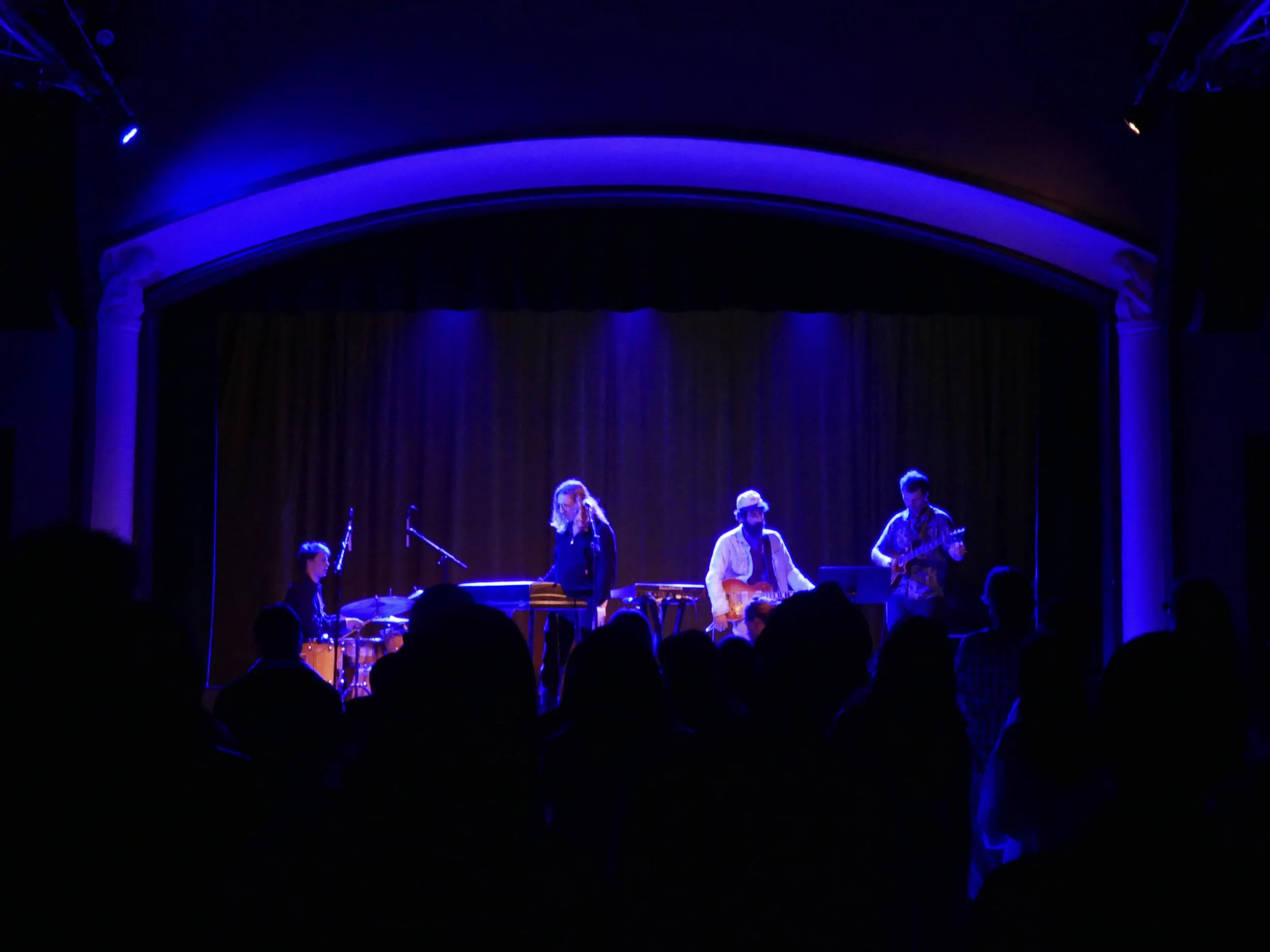 A live musical performance on stage with band members playing guitars and drums, illuminated by blue stage lighting, with an audience in the foreground.