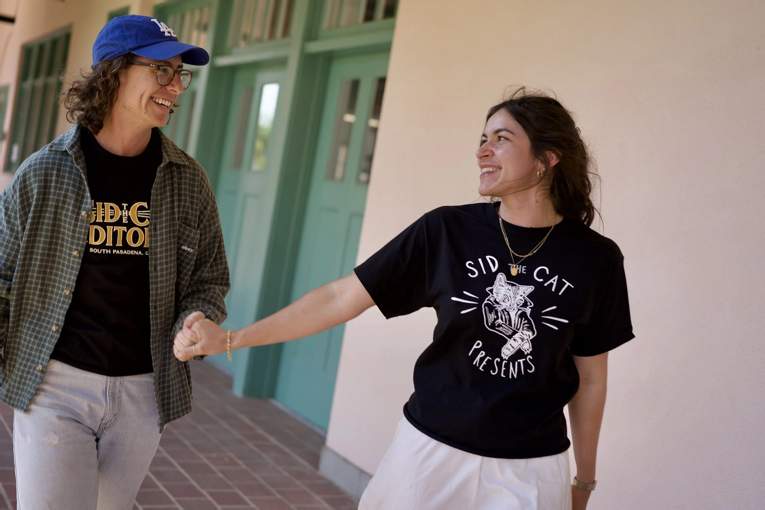 Two women shaking hands and smiling at each other outside a building with green doors and windows.