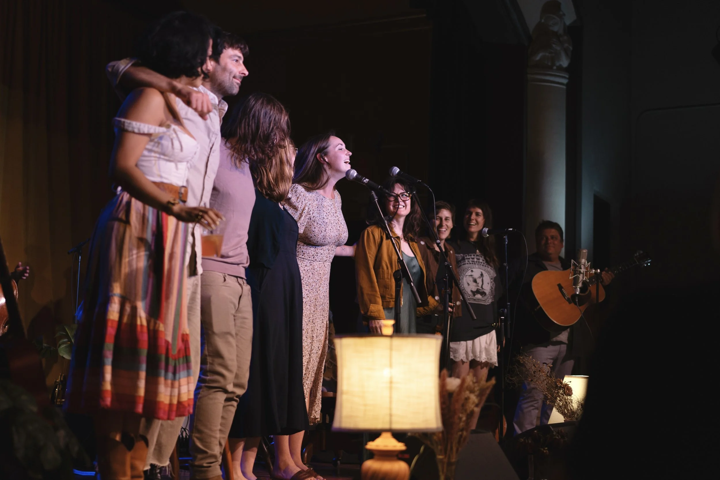 A group of people standing on a stage, some with microphones and one playing a guitar, in a warmly lit indoor setting, possibly during a performance or event.
