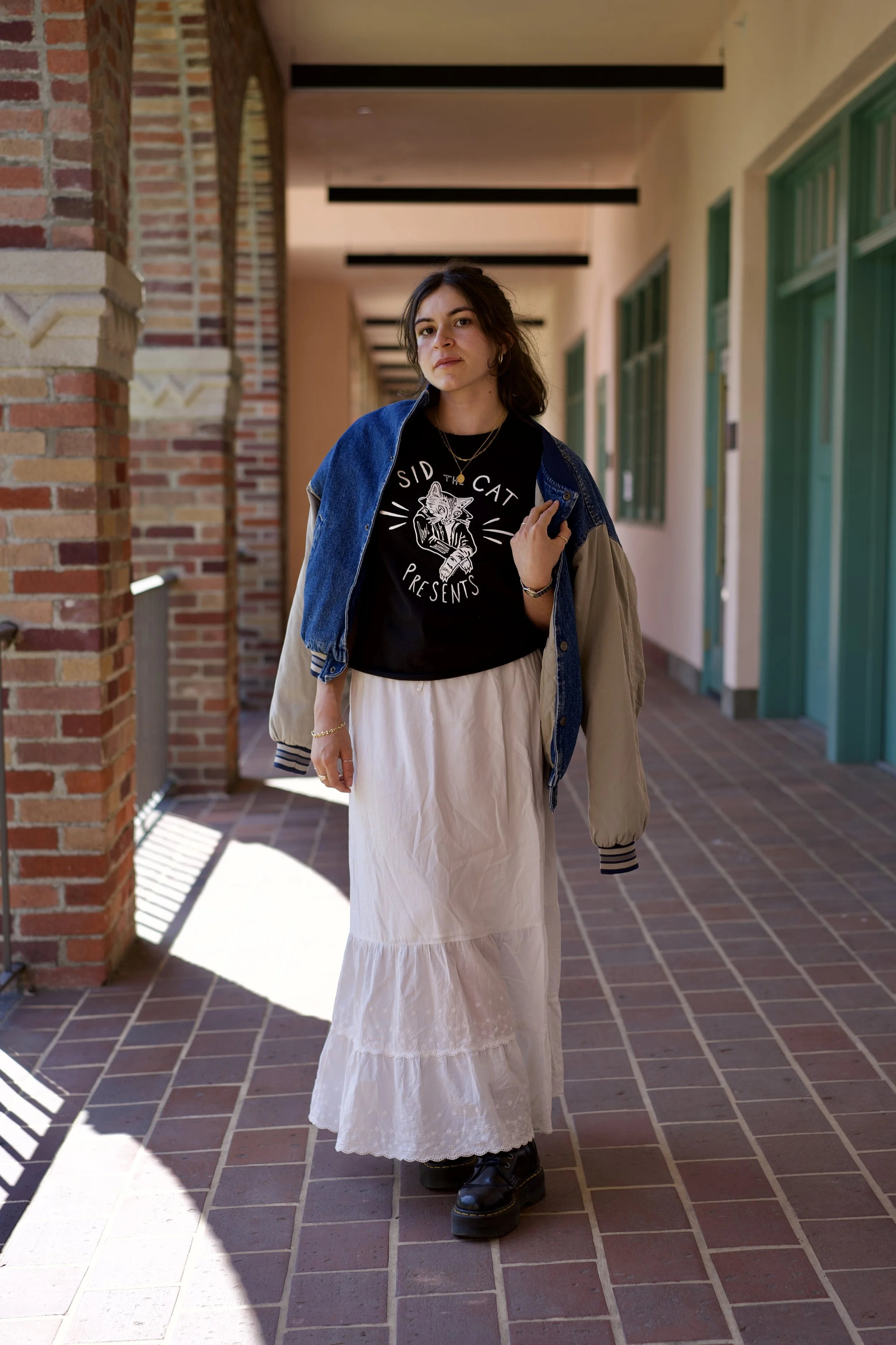 Young woman walking in a corridor with brick walls, wearing a black graphic t-shirt, a beige and denim jacket, long white skirt, and black boots, holding the jacket with one hand and looking into the camera.