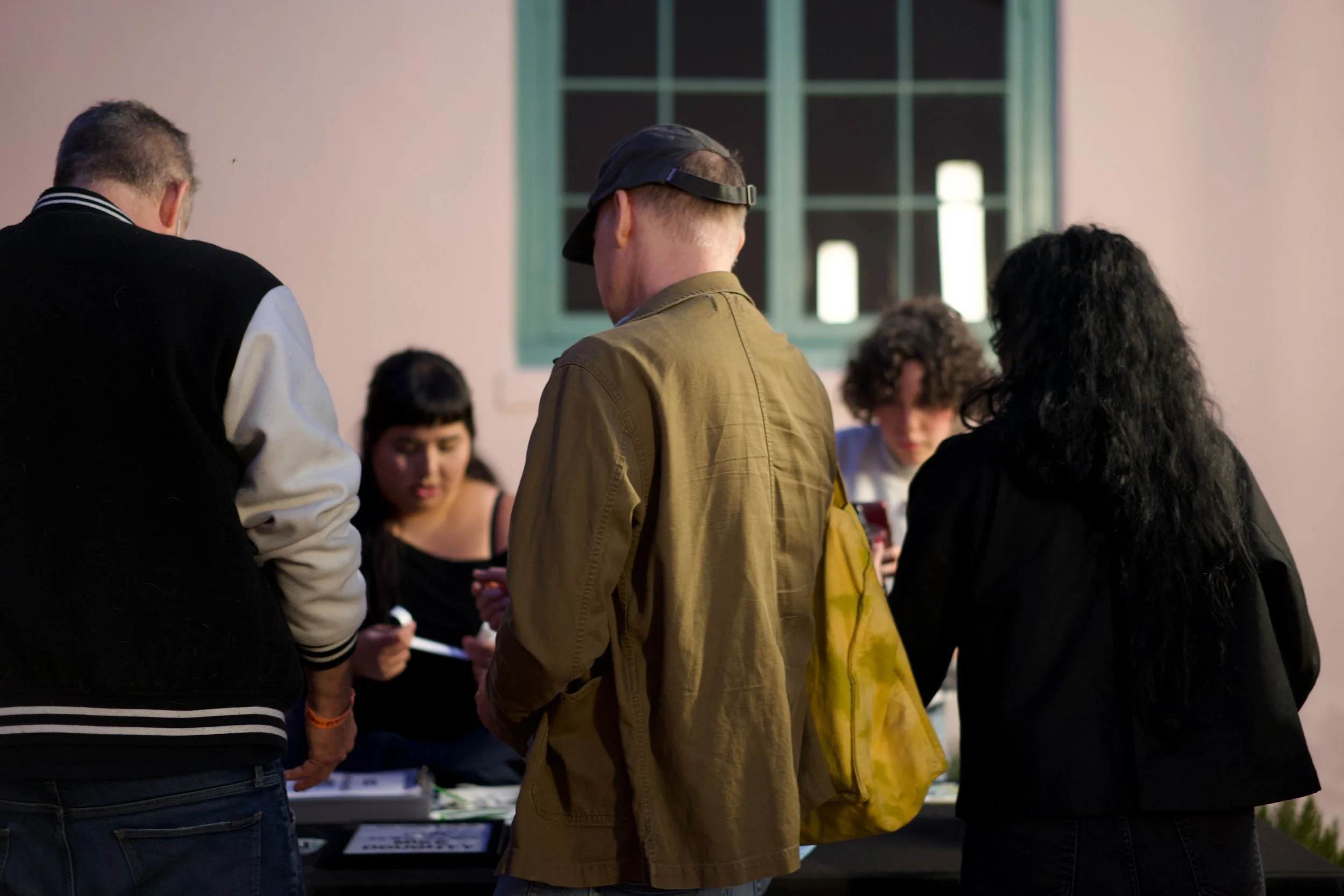 People gathered around a table at an indoor event, looking at items or information displayed on the table, in front of a window with green framing.