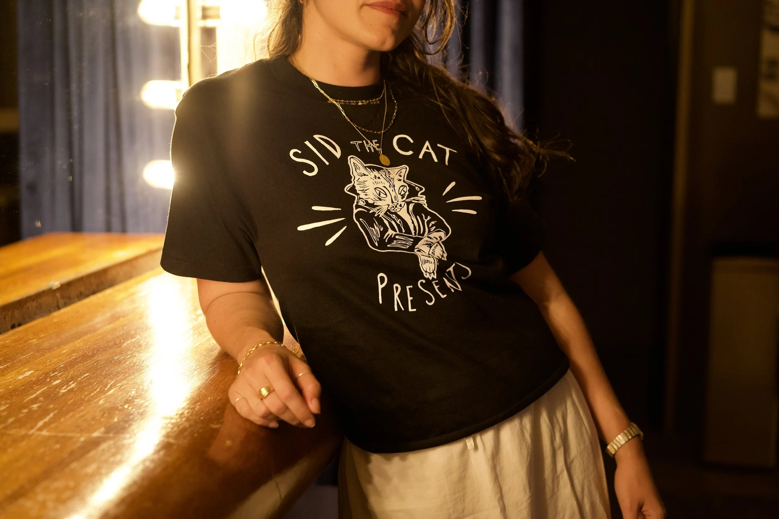 Person wearing a black T-shirt with a graphic of a cat and the text "Sidd The Cat" against a dimly lit background with string lights.