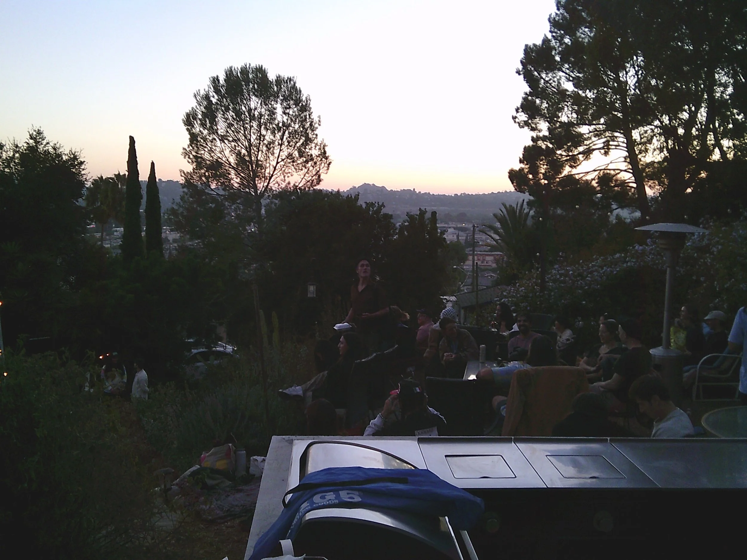 A group of people gathered outdoors during dusk, with trees and hills in the background. Some are seated, engaging in conversation or looking around.