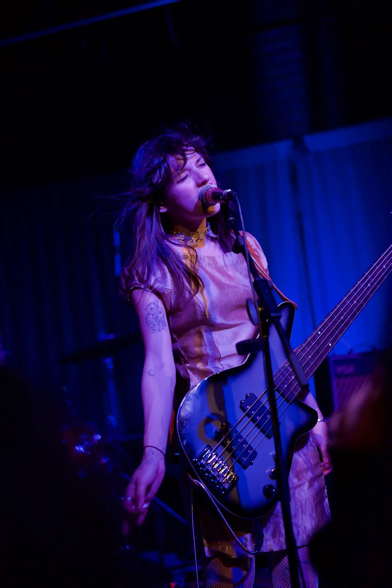 Female musician singing into a microphone and playing an electric guitar on stage with blue and purple lighting, wearing a pink satin top and fishnet pants.