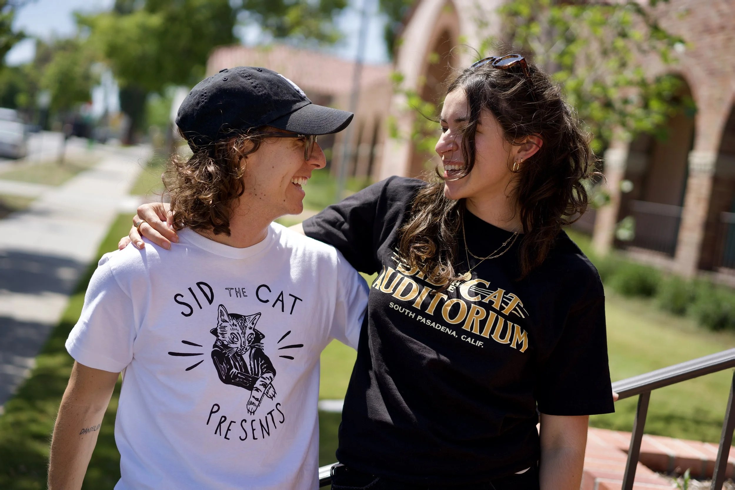 Two women smiling at each other outdoors on a sunny day, one wearing a black cap and glasses, the other with sunglasses on her head, walking along a sidewalk with trees and brick buildings in the background.