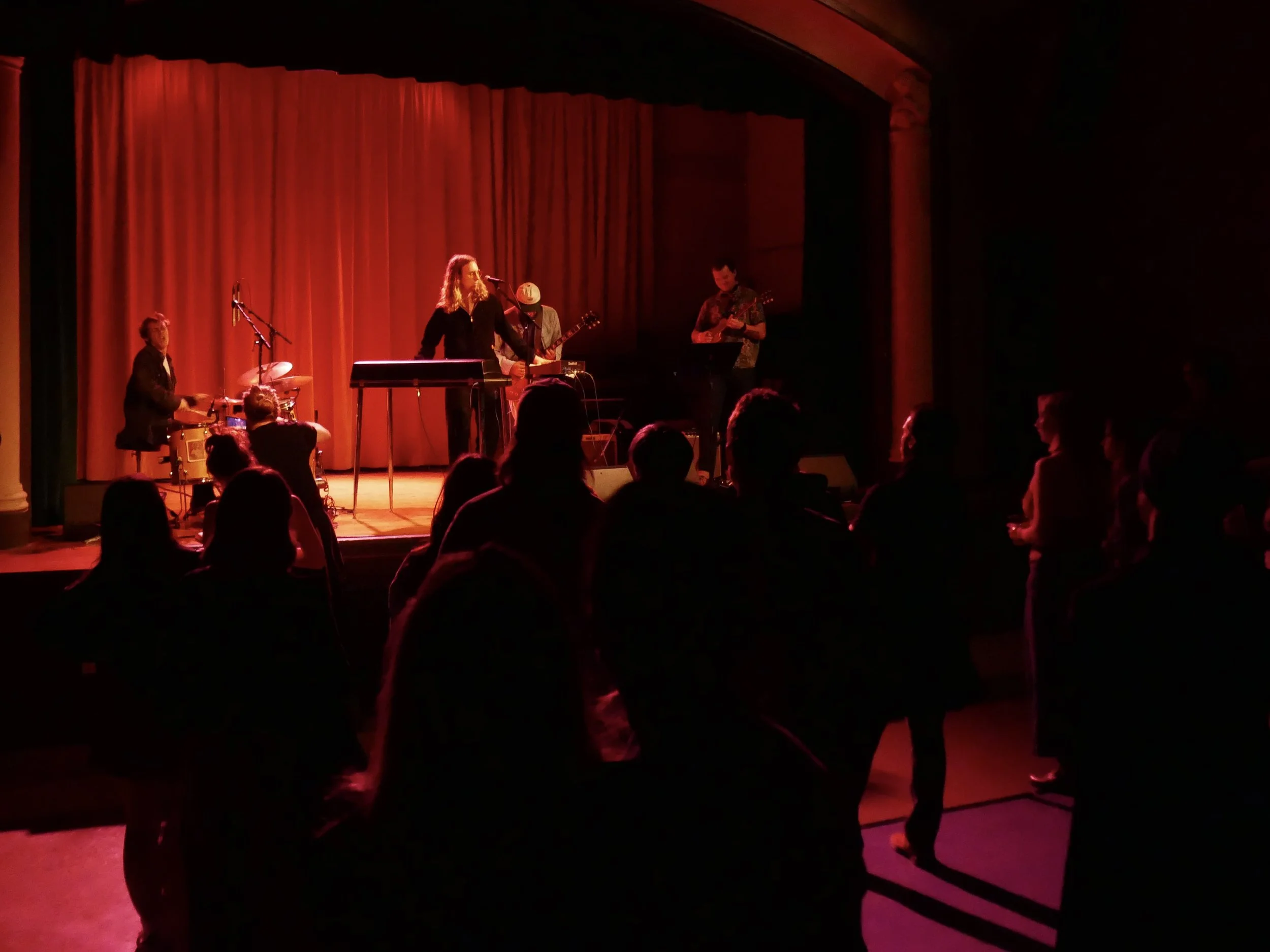 A band performing on stage with a red curtain backdrop, including a drummer, a keyboardist, and guitarists, in a dimly lit venue with audience silhouettes in the foreground.