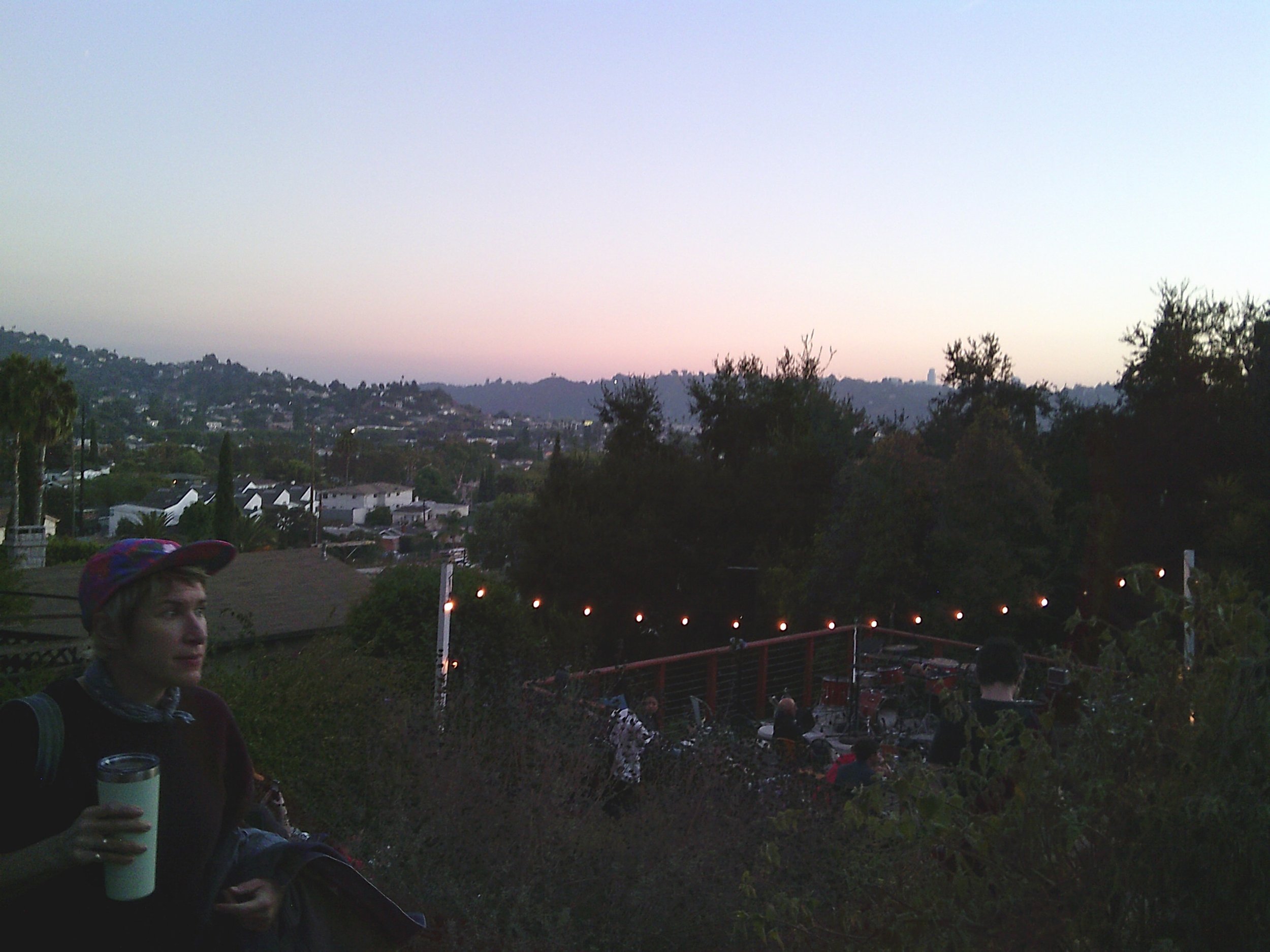 People gathered outdoors during evening sunset, with trees, houses, and distant hills in the background.